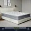 The image features a white bed with a grey comforter and a grey pillow. The bed is a queen size and has a mattress with a plush medium firmness. The bed is positioned in a bedroom with a white carpet. There is also a nightstand next to the bed.