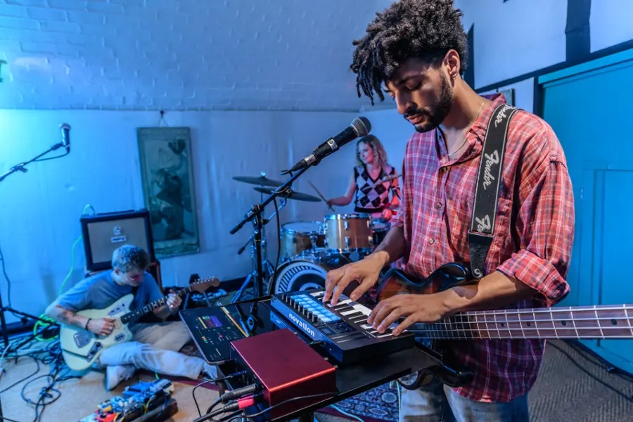 The image shows a man playing a keyboard, with a guitarist and drummer behind him. The man is playing a Novation keyboard, while the guitarist is using a Fender guitar. The drummer is positioned between the keyboard player and the guitarist, creating a dynamic and lively musical performance.