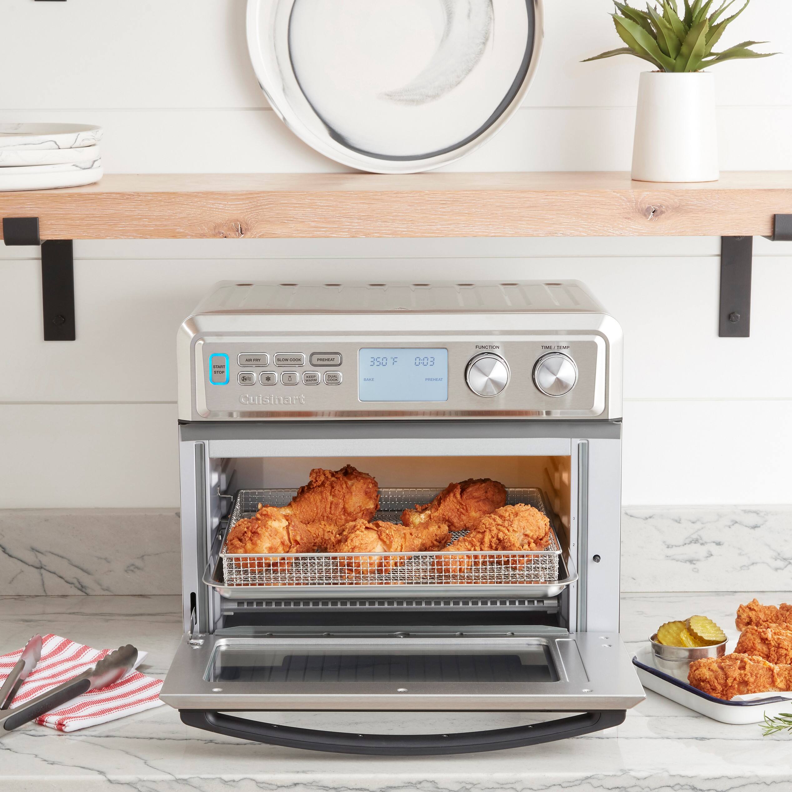 The image shows a Cuisinart toaster oven with a tray of chicken inside. The oven is placed on a counter, and there are utensils nearby, including a knife and a fork. Additionally, there is a potted plant on the counter, and a bowl can be seen in the background.