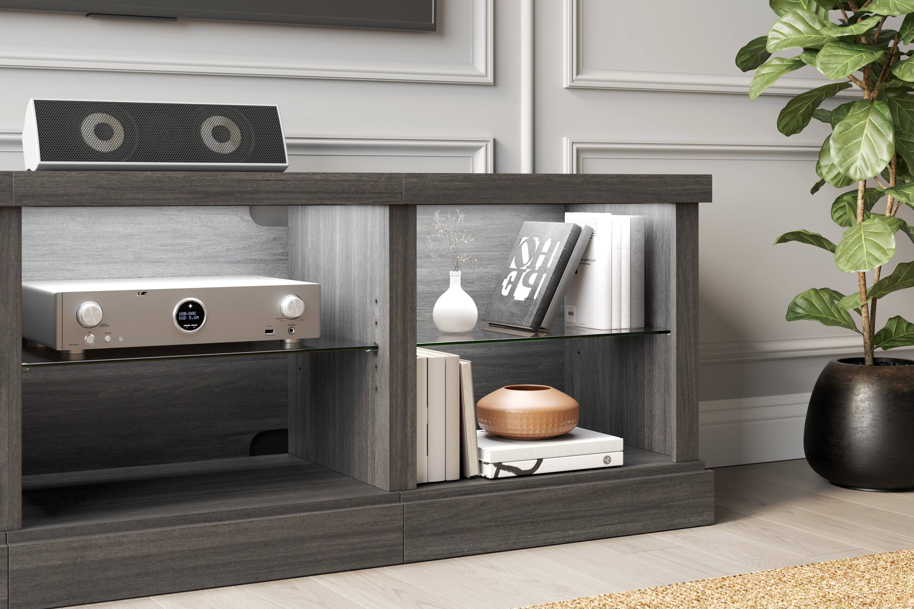 The image shows a wooden entertainment center with a speaker, a vase, and a potted plant. The speaker is located on the left side of the center, while the vase is positioned in the middle. The potted plant is situated on the right side of the center. The entertainment center is placed on a wooden floor, and there is a bookshelf with books on it. The arrangement of the items creates a cozy and inviting atmosphere in the room.