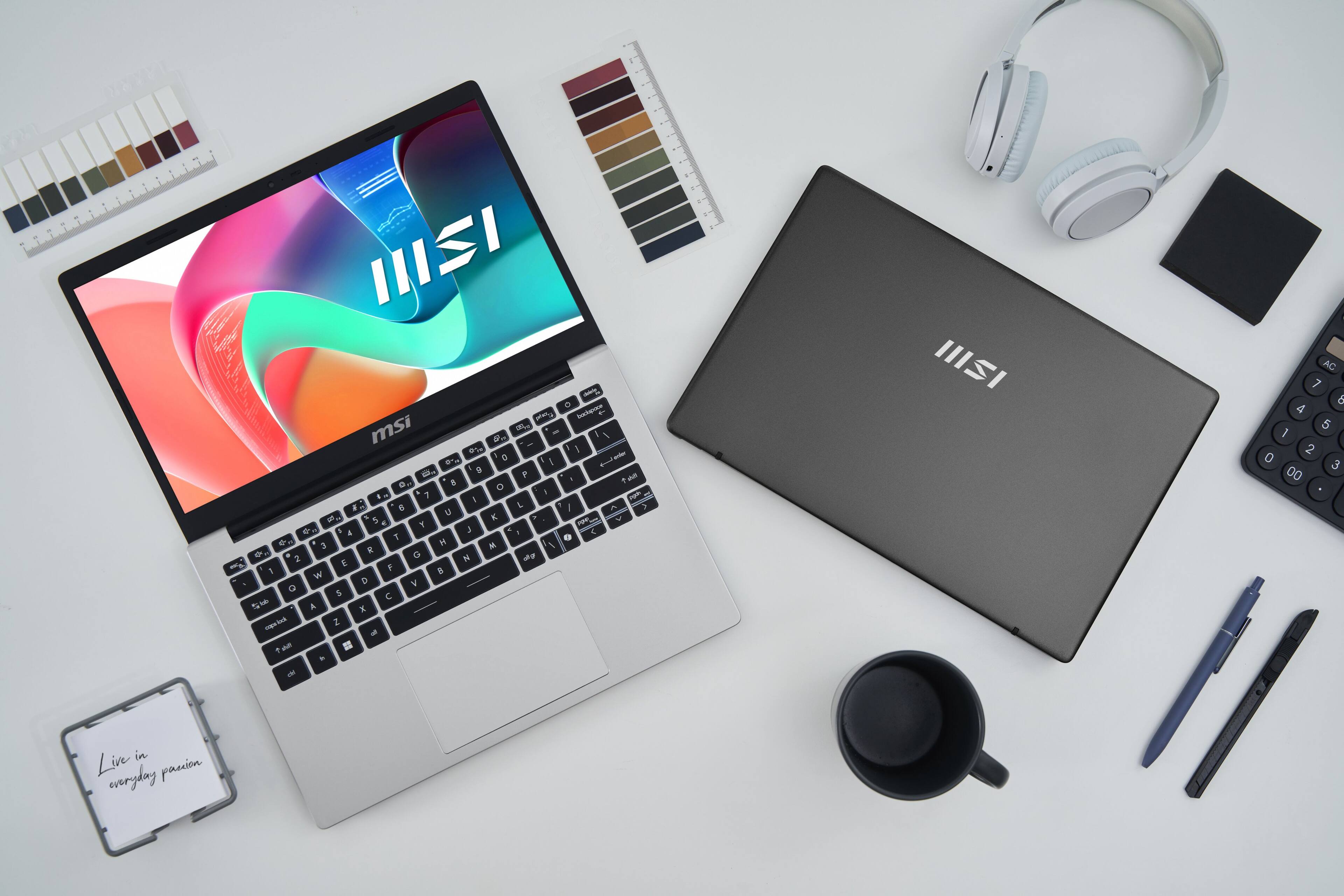 The image features a white table with two laptops, one of which is an MSI laptop. There are also headphones, a cup, and a pen on the table. The table is covered with various items, including a laptop, headphones, a cup, a pen, and a book. The scene suggests that someone is working or studying, utilizing the laptops and other items to complete their tasks.