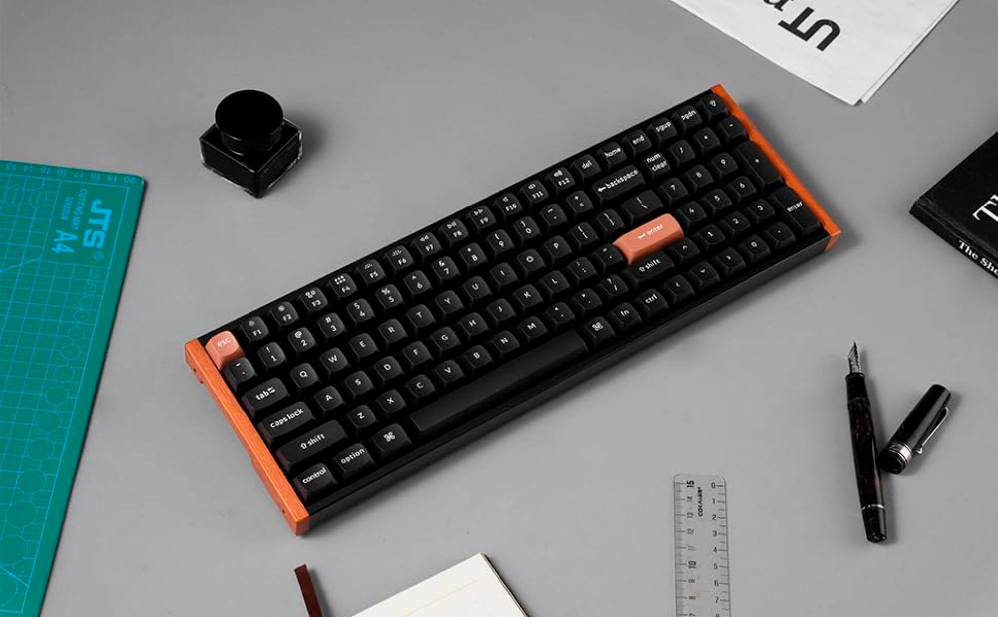 The image shows a keyboard placed on a table, surrounded by various items. The keyboard has a unique design, featuring orange and black keys. The table also has a ruler, a book, and a pair of scissors. The keyboard is positioned in the center of the table, with the ruler to the right and the book to the left. The scissors are located at the top right corner of the table.