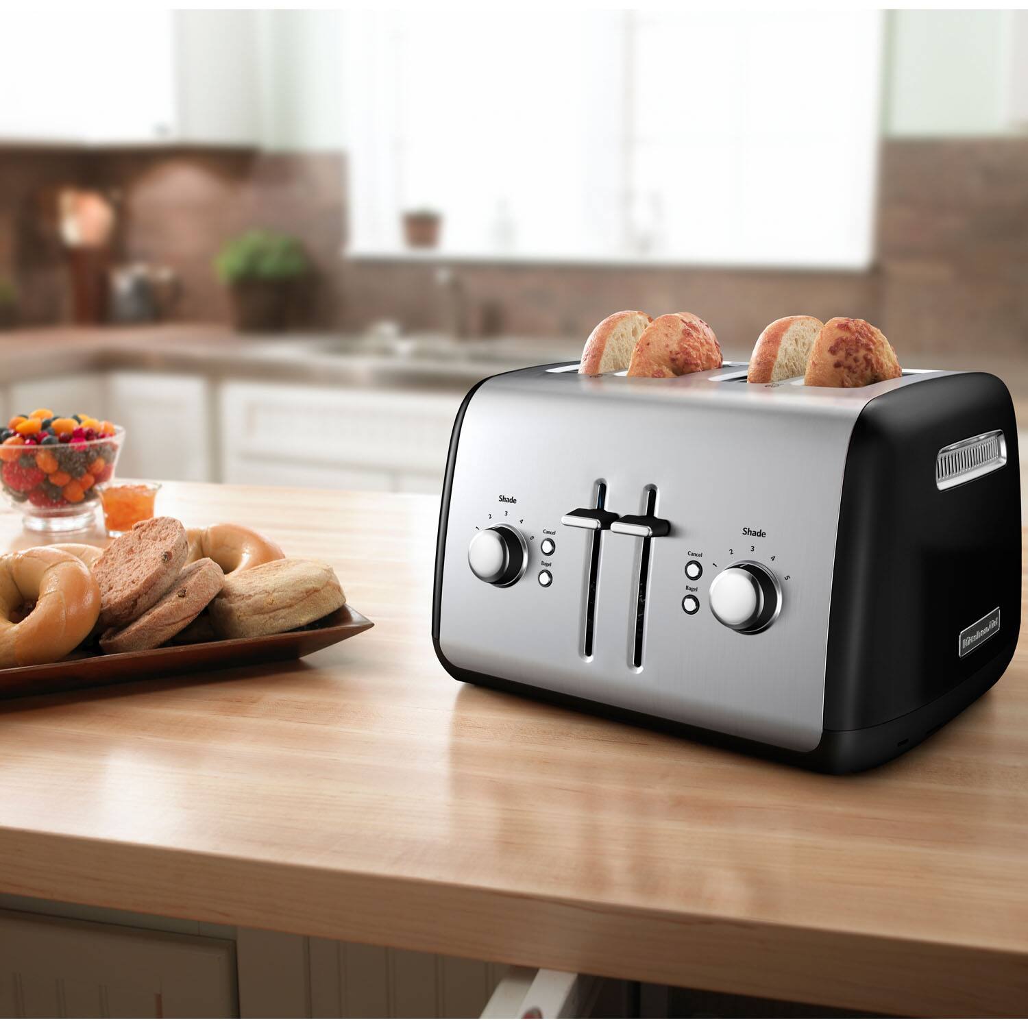 The image shows a toaster oven on a countertop with a tray of bagels and a bowl of fruit. The toaster oven has a timer and a knob to control the heat. The countertop also has a sink nearby.