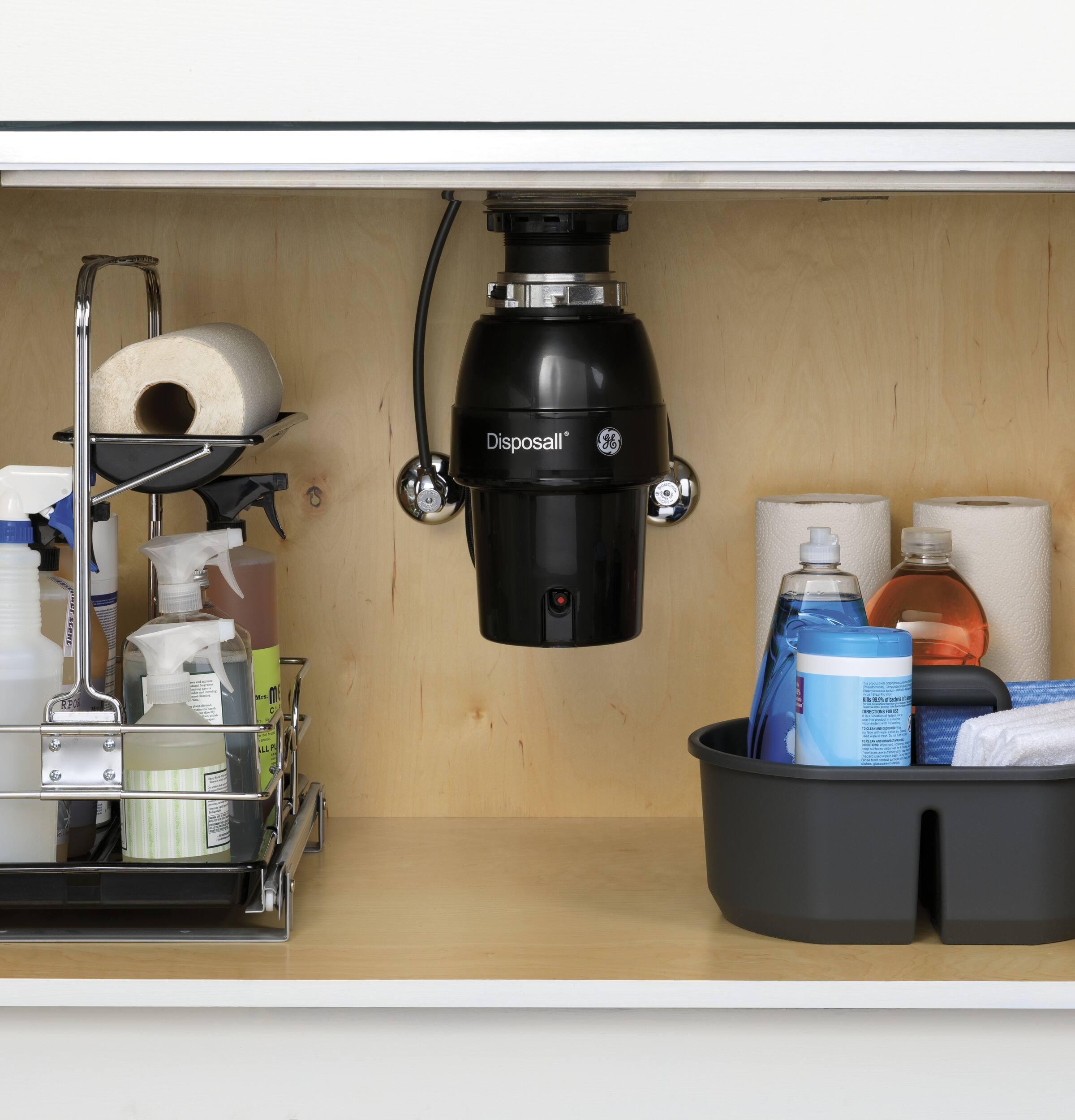 The image shows a bathroom counter with a black garbage disposal unit, a black bucket, and various cleaning supplies. The counter is made of wood and has a shelf with a variety of items on it. There are several bottles of different sizes, some of which are placed in a bucket. Additionally, there are two toilet paper rolls on the counter. The scene suggests a well-organized and functional bathroom space.