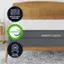 The image features a wooden bed frame with a mattress on top of it. The mattress is made in the USA and contains CertiPUR-US foam, which is a certified flexible polyurethane foam. The mattress is also certified by CertiPUR-US, indicating that it meets their standards for safety and quality. The bed frame is part of the Ghostbed Luxe collection, which comes with a 25-year warranty.