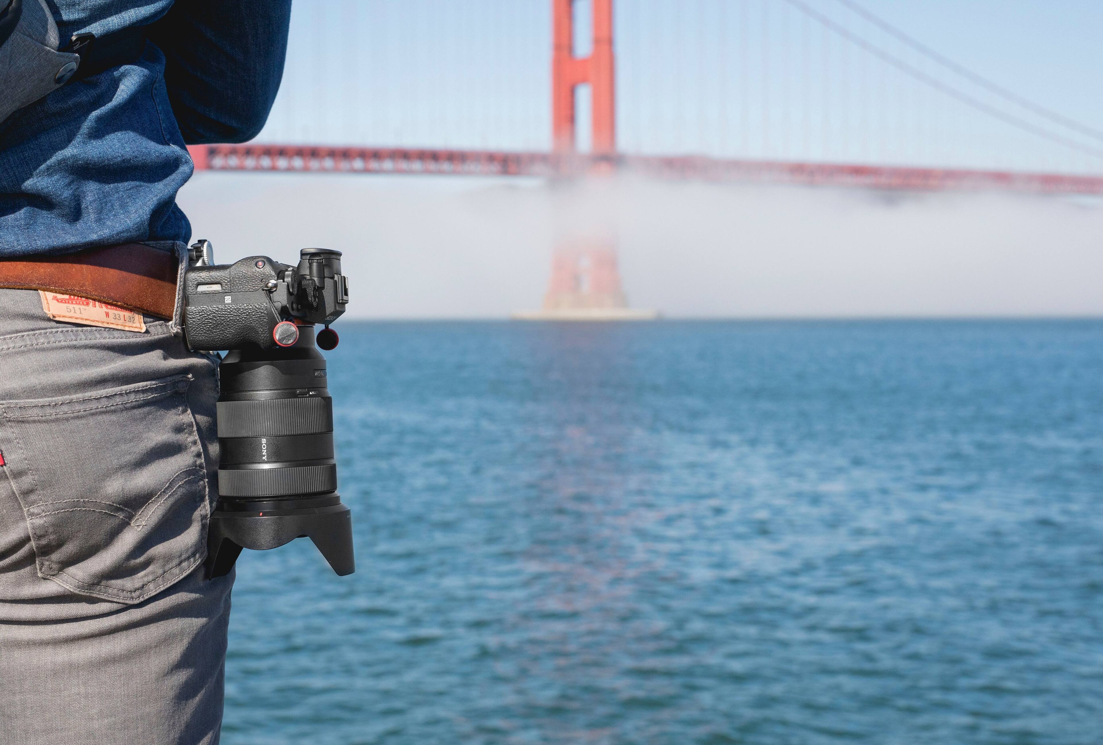 The text "SEO b11 - 1 33 L2T - EL" is not relevant to the image. The image shows a person wearing a blue shirt and jeans, holding a camera and standing near a body of water, possibly the ocean. The person is also wearing a brown belt.