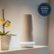 The image features a white device, possibly a security system, sitting on a shelf next to a vase with flowers. The device is prominently displayed in the scene, and the vase with flowers adds a touch of elegance to the setting. The image is categorized as Household_Detectors_and_Sensors, and it requires a SimpliSafe Security System.