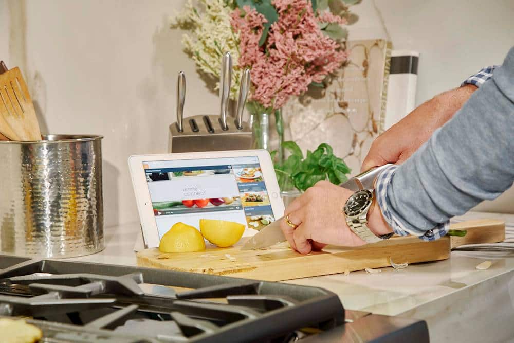 I'm sorry, but I cannot group or correct the text "Hore connect O" as it does not make sense or provide any context. The image shows a person using a tablet in a kitchen, surrounded by various kitchen items such as a cutting board, knife, and a vase of flowers. The tablet is displaying a recipe, and the person is preparing food.