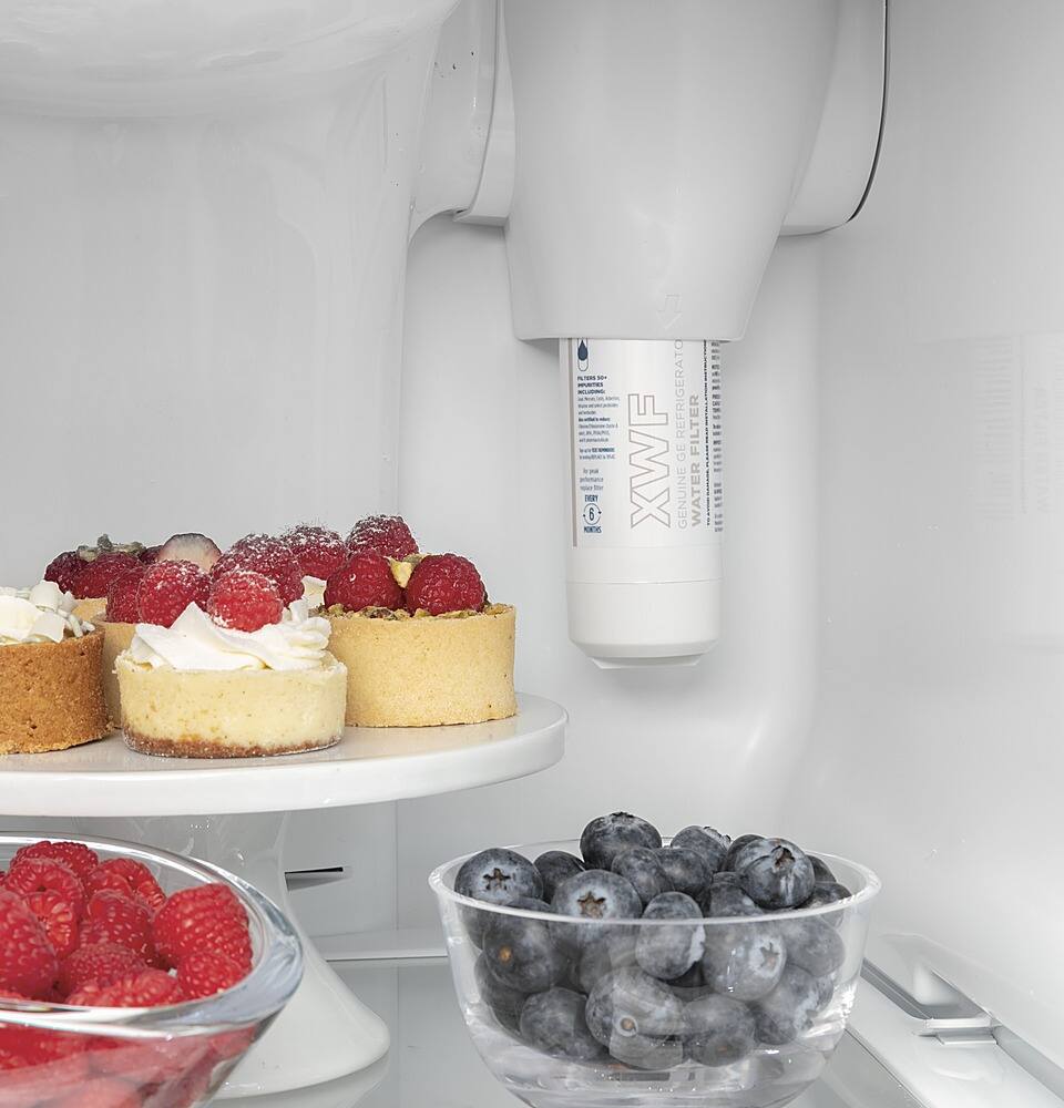 The image shows a refrigerator with a water dispenser on the door. The refrigerator is filled with various food items, including a bowl of berries, a cake, and a bowl of fruit. There are also several cakes placed inside the refrigerator. The refrigerator is a large, white appliance that is well-stocked with food and drinks.
