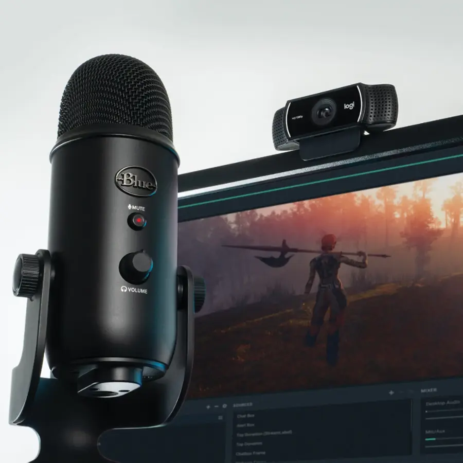 The image features a Blue Yeti microphone sitting on top of a computer monitor. The microphone is positioned in front of the monitor, and a webcam is also present in the scene. The monitor displays a video game, possibly a stream or a chat session. The setup suggests that the user is engaged in a video game or streaming session, using the microphone and webcam to communicate with others or record their gameplay.