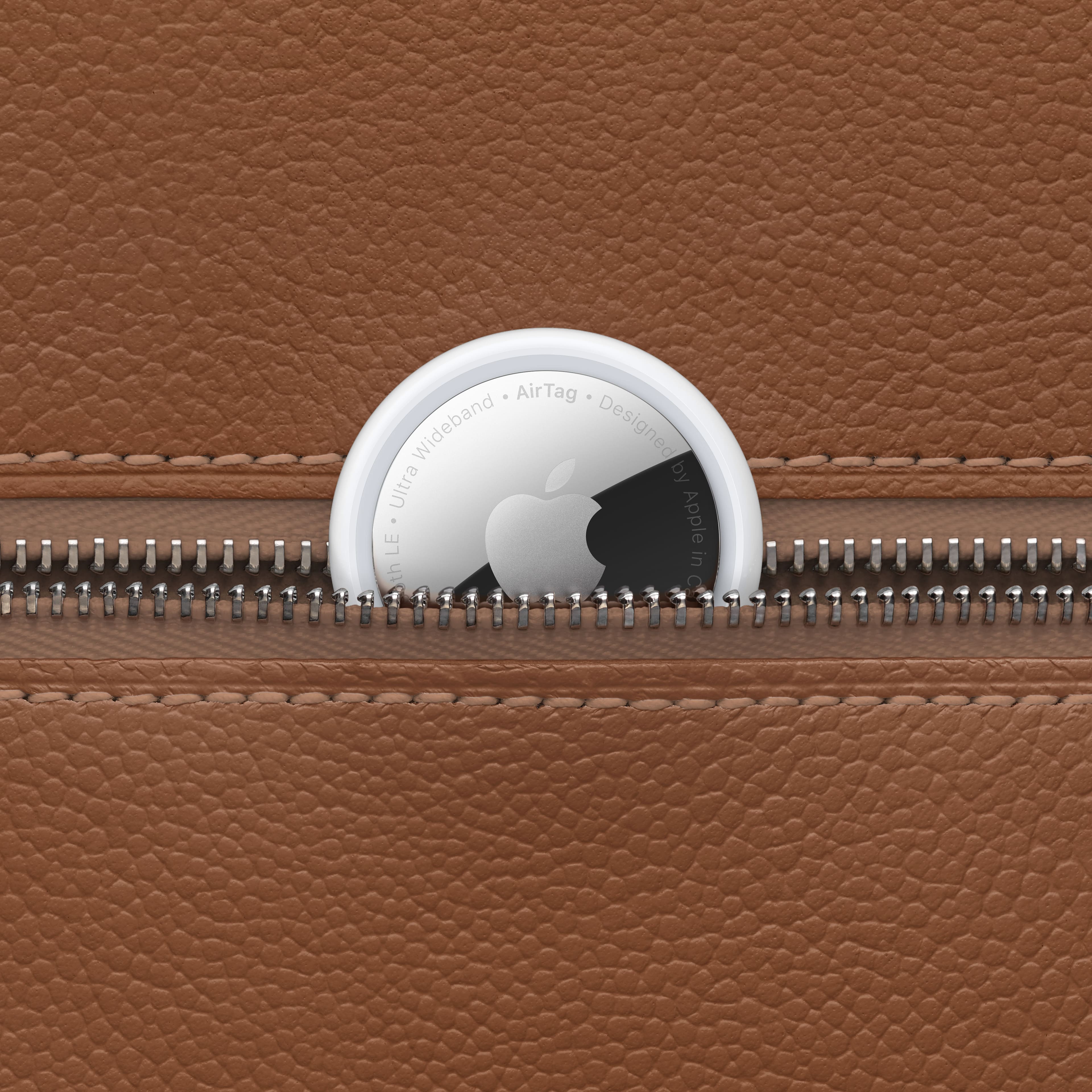 The image features a coin-sized Apple AirTag attached to a brown leather wallet. The AirTag is designed by Ultra LE + Apple, and it is a wideband tracker. The wallet is zipped up, and the AirTag is visible through the zipper.