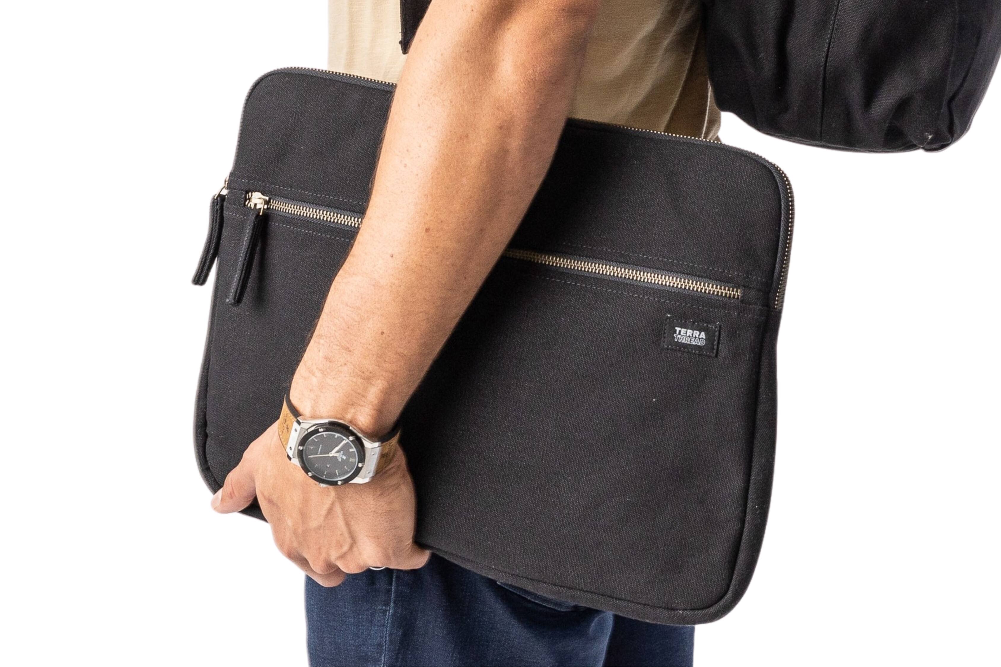 The image shows a person holding a black laptop sleeve with a zipper and a small pocket on the front. The person is also wearing a watch on their wrist.
