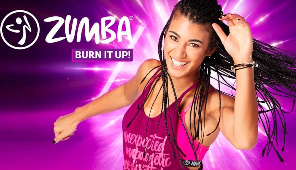 The image features a woman wearing a purple shirt and braids, who is smiling and dancing. She is the main focus of the image, and her expression and body language suggest that she is enjoying herself. The background of the image is purple, which adds to the overall color scheme of the scene. The woman's attire and the purple background create a vibrant and energetic atmosphere, which is likely related to the Zumba brand and its focus on fitness and dance.