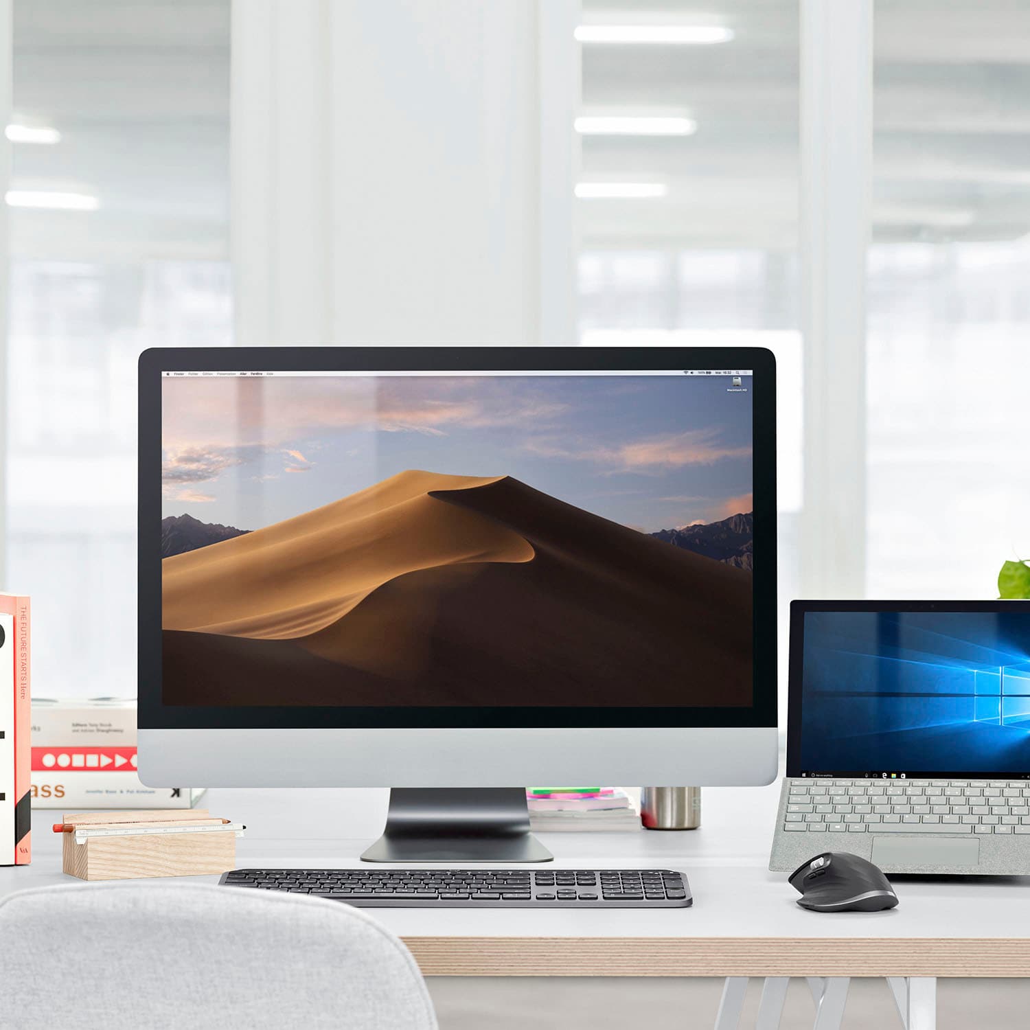 The image shows a desk with a desktop computer, a laptop, a keyboard, and a mouse. There is also a book and a potted plant on the desk. The desk is located in front of a window, which provides natural light to the workspace. The setup appears to be a comfortable and functional workstation for various tasks, such as work, study, or entertainment.