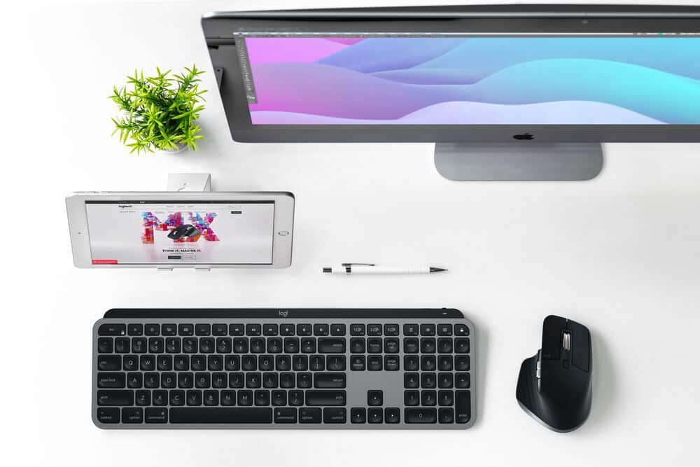The image features a computer setup with a keyboard, mouse, and monitor. The keyboard is placed in front of the monitor, and the mouse is located to the right of the keyboard. There is also a cell phone placed next to the keyboard. The monitor is turned on, displaying a colorful screen. The arrangement of these items suggests a workspace or a home office setup.