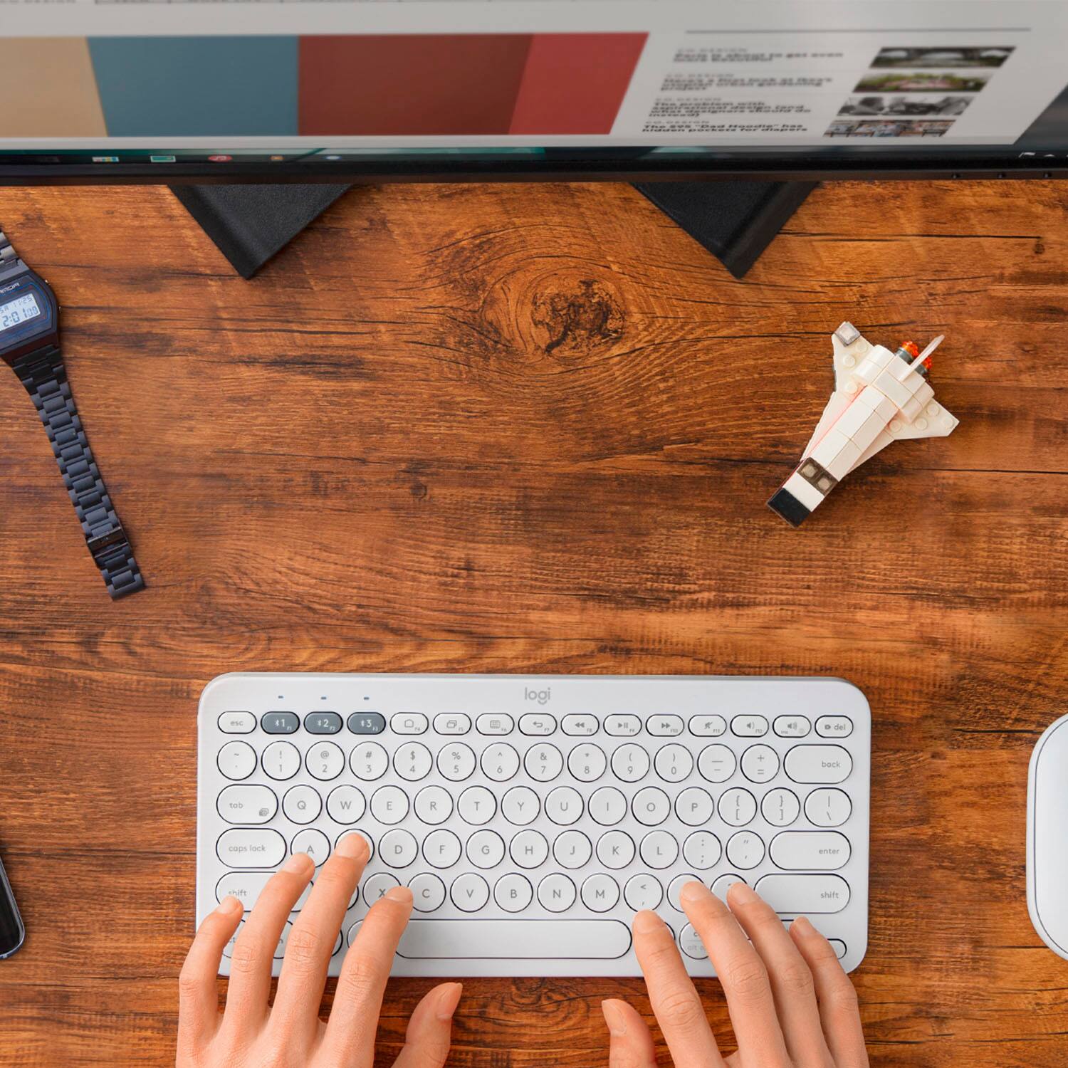 The image shows a person typing on a white keyboard. The keyboard has a unique design, with the letters and numbers arranged in a different order than the standard QWERTY layout. The person's hands are visible on the keyboard, and they are typing with precision. The scene is set on a wooden desk, which also has a cell phone and a clock nearby.