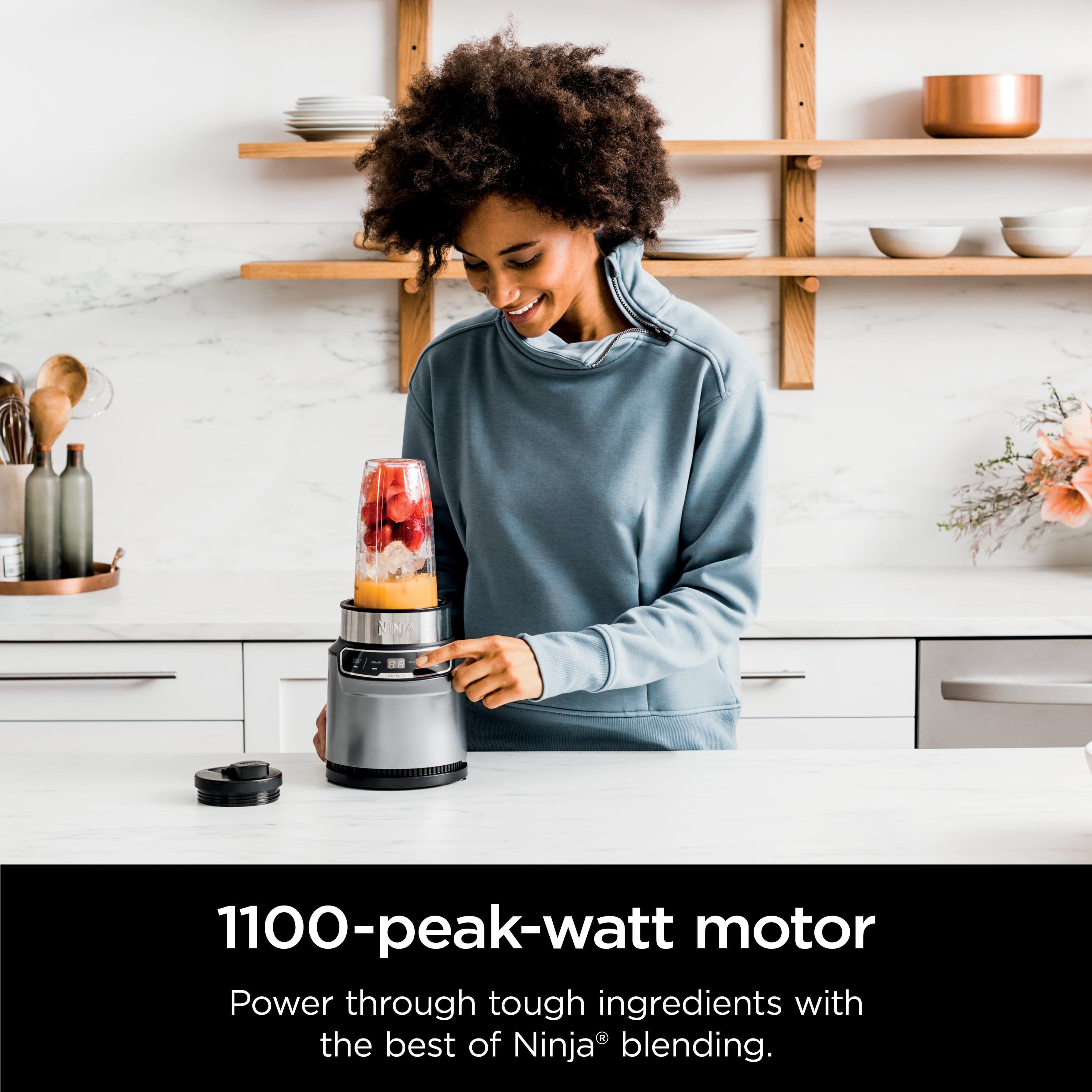 The image features a woman standing in a kitchen, smiling and holding a blender with a mixture inside. The blender is placed on a countertop, and there are several bowls and cups nearby. The woman appears to be enjoying her time in the kitchen. The image is promoting a 1100-peak-watt motor that can power through tough ingredients with the best of Ninja blending.