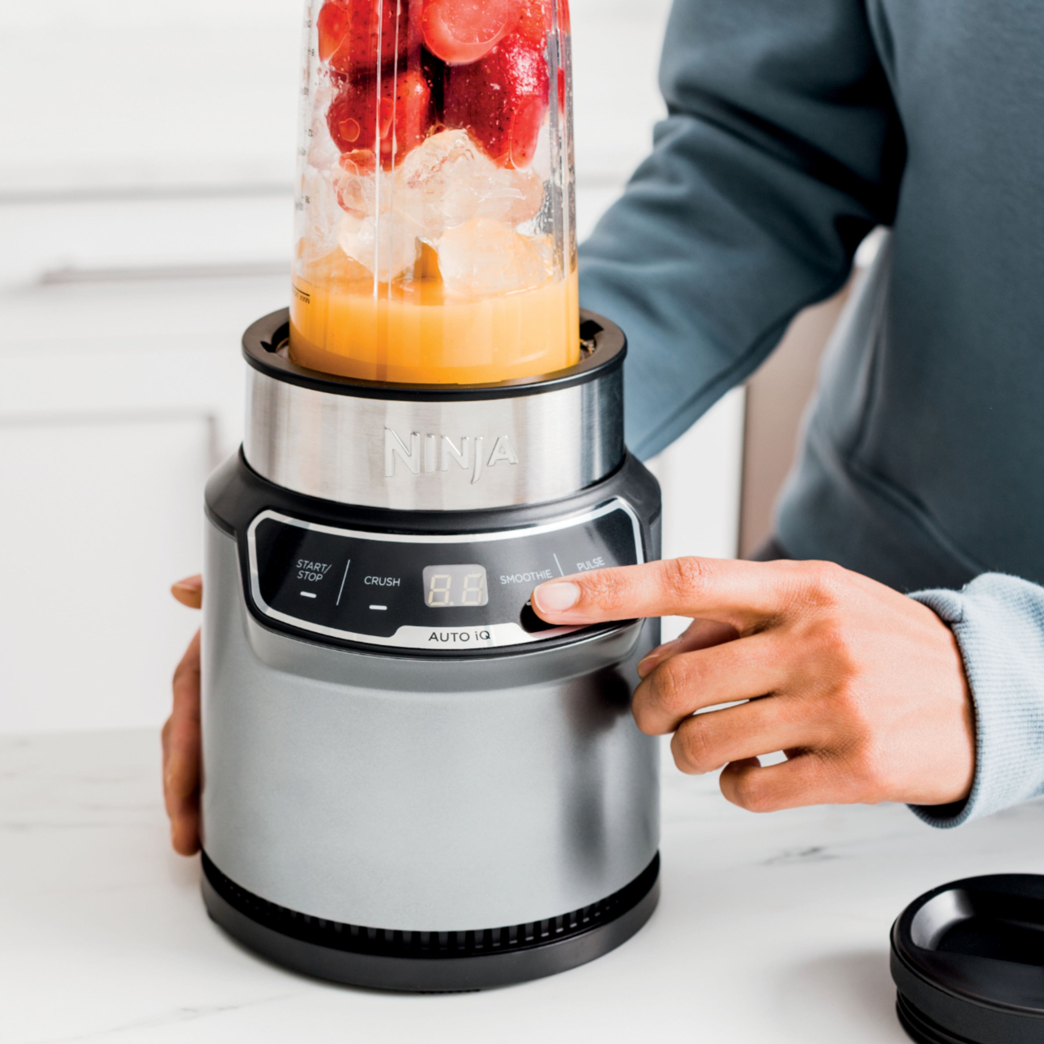 The image features a person using a Ninja blender to make a smoothie. The blender is filled with strawberries and ice, and the person is pressing a button on the blender. The image also showcases the Auto-iQ Technology, which offers timed intelligent blending programs that do the work for you, eliminating the need for guesswork.