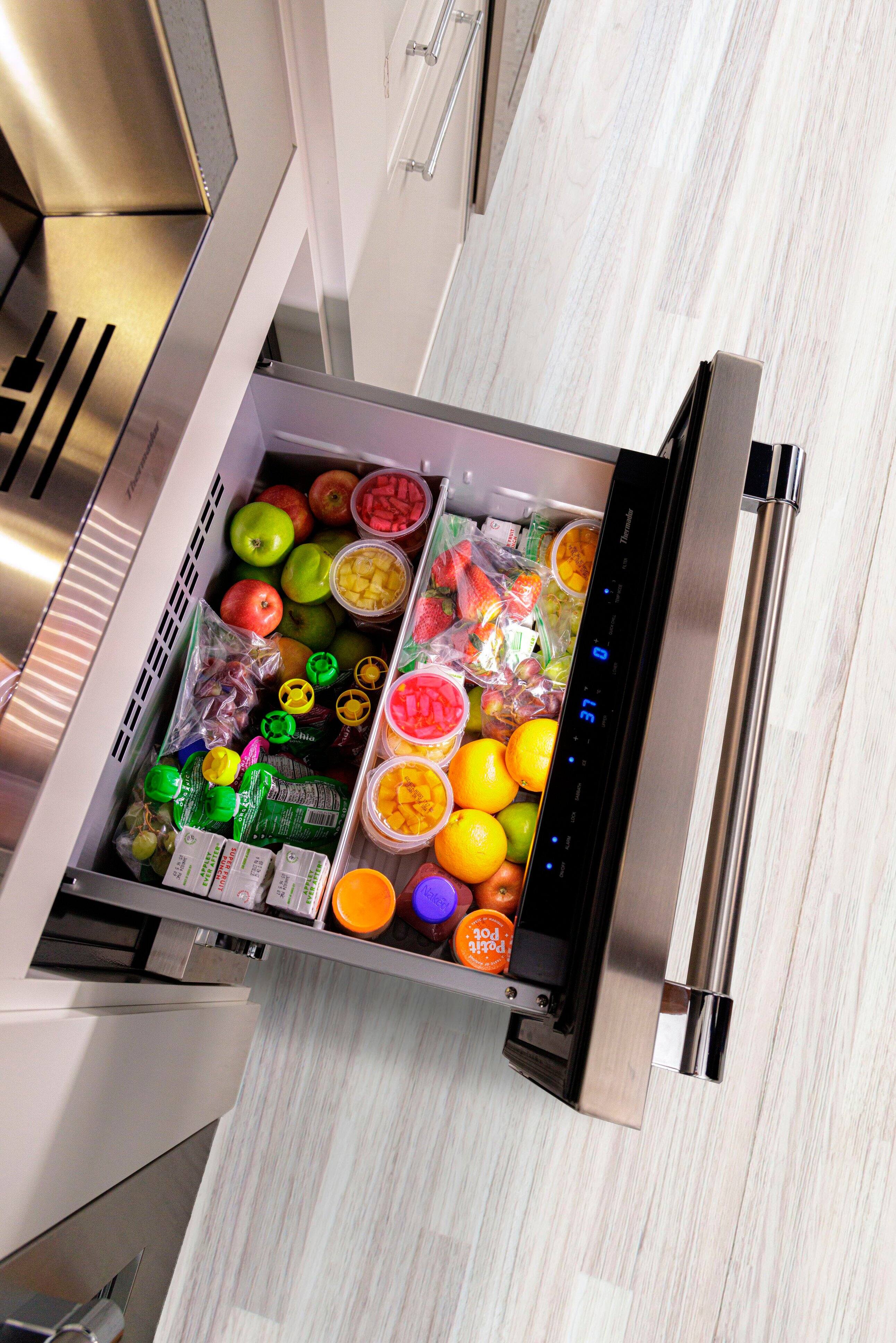 The image shows a Thermador refrigerator with its door open, revealing a variety of food items inside. There are several apples and oranges placed in different compartments, as well as a bowl of fruit. The refrigerator is filled with a diverse assortment of food, making it an appealing and well-stocked appliance.