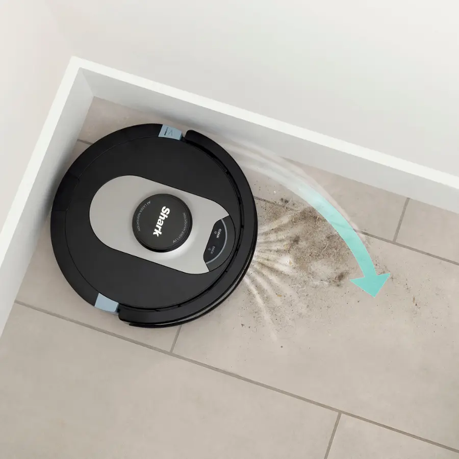 The image features a woman sitting on a couch in a living room, holding a cell phone. In front of her, there is a robotic vacuum cleaner, which is an example of a bagless self-empty base. This type of vacuum cleaner is unique because it empties itself into a base that holds up to 60 days of debris, eliminating the need for frequent bag replacement or manual emptying. The woman appears to be using the cell phone, possibly controlling the robotic vacuum cleaner or browsing the internet.