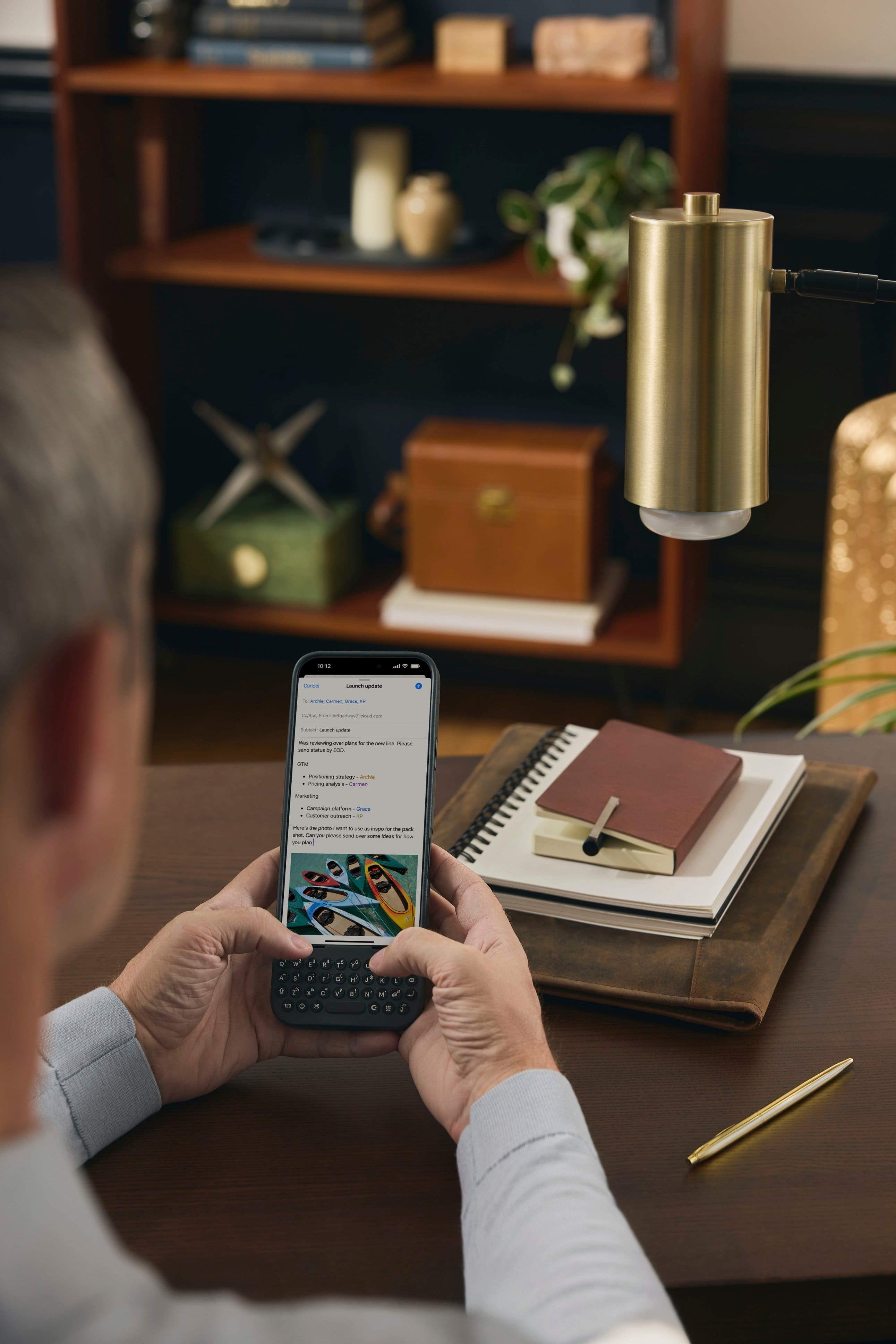 The man is sitting at a desk and looking at his cell phone. He is holding the phone in his hands, possibly texting or browsing the internet. The desk is cluttered with various items, including a book, a vase, and a potted plant. There is also a remote control on the desk, and a clock is visible in the background. The man appears to be focused on his phone, possibly checking his messages or working on a task.