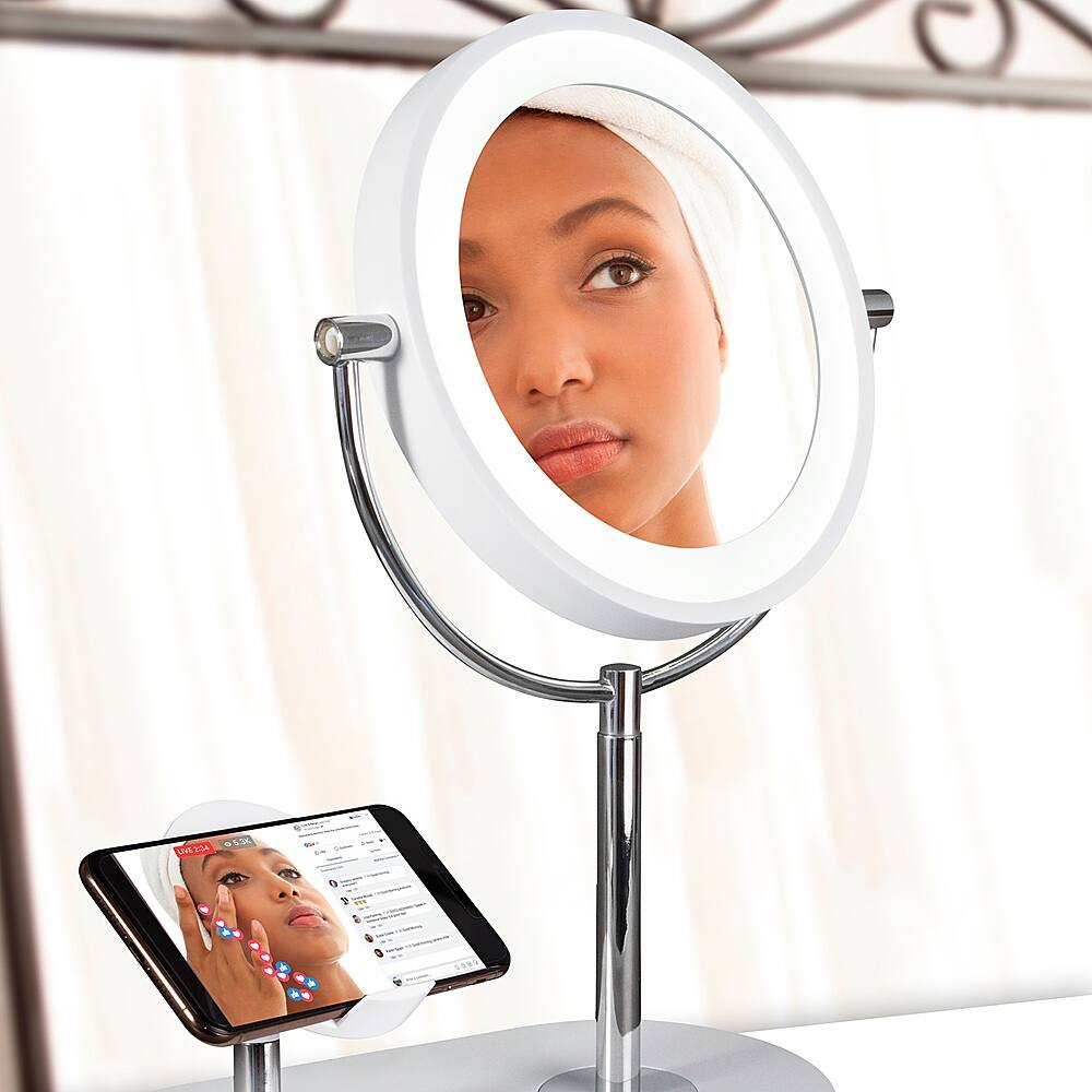 The image shows a woman looking at herself in a mirror, with a cell phone placed nearby. The mirror is positioned on a stand, and the woman appears to be using the cell phone to take a picture of herself or view her reflection. The scene suggests that she is capturing a selfie or checking her appearance on the phone.