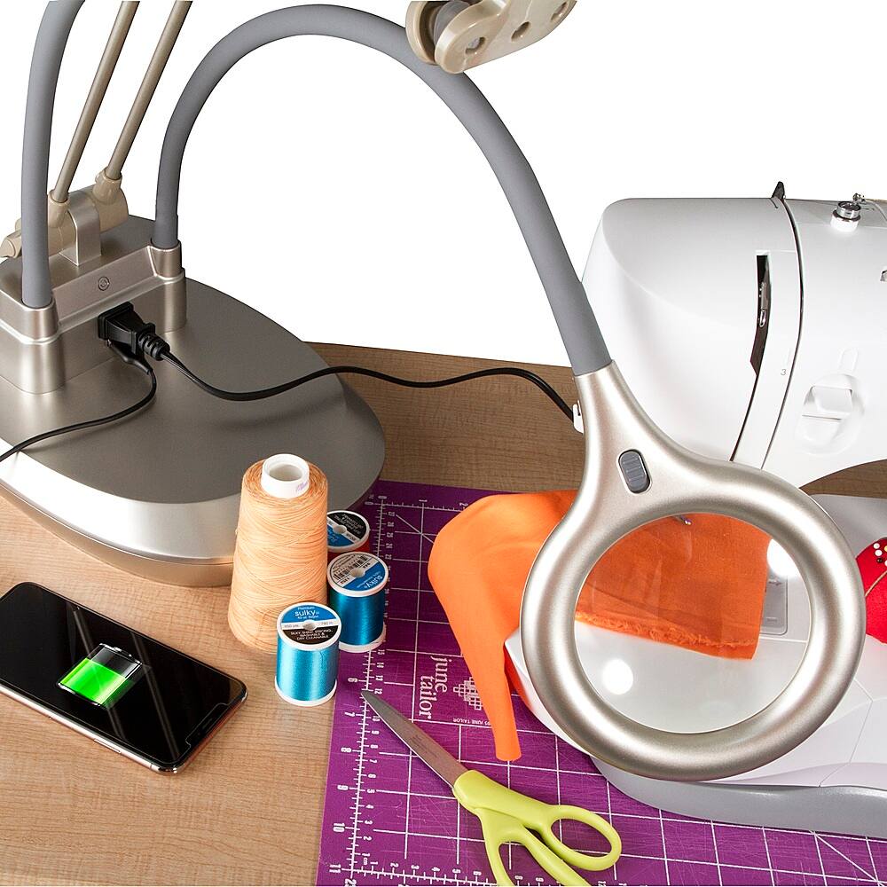 The image shows a sewing table with various sewing tools and materials. There is a pair of scissors, a sewing machine, a cell phone, and a spool of thread. The table also has a measuring tape and a pair of pins. The sewing machine is on the left side of the table, while the cell phone is on the right side. The scissors are located near the center of the table, and the spool of thread is in the middle. The measuring tape is placed on the right side of the table, and the pair of pins is located near the bottom right corner.