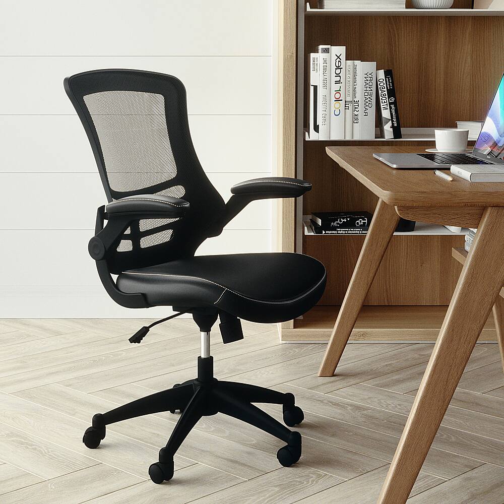 The image features a black office chair with wheels, positioned in front of a wooden desk. The desk is cluttered with various items, including several books, a laptop, a cup, and a bowl. The books are scattered around the desk, with some placed closer to the laptop and others further away. The cup and bowl are situated near the edge of the desk. The office chair is placed in front of the desk, ready for use.