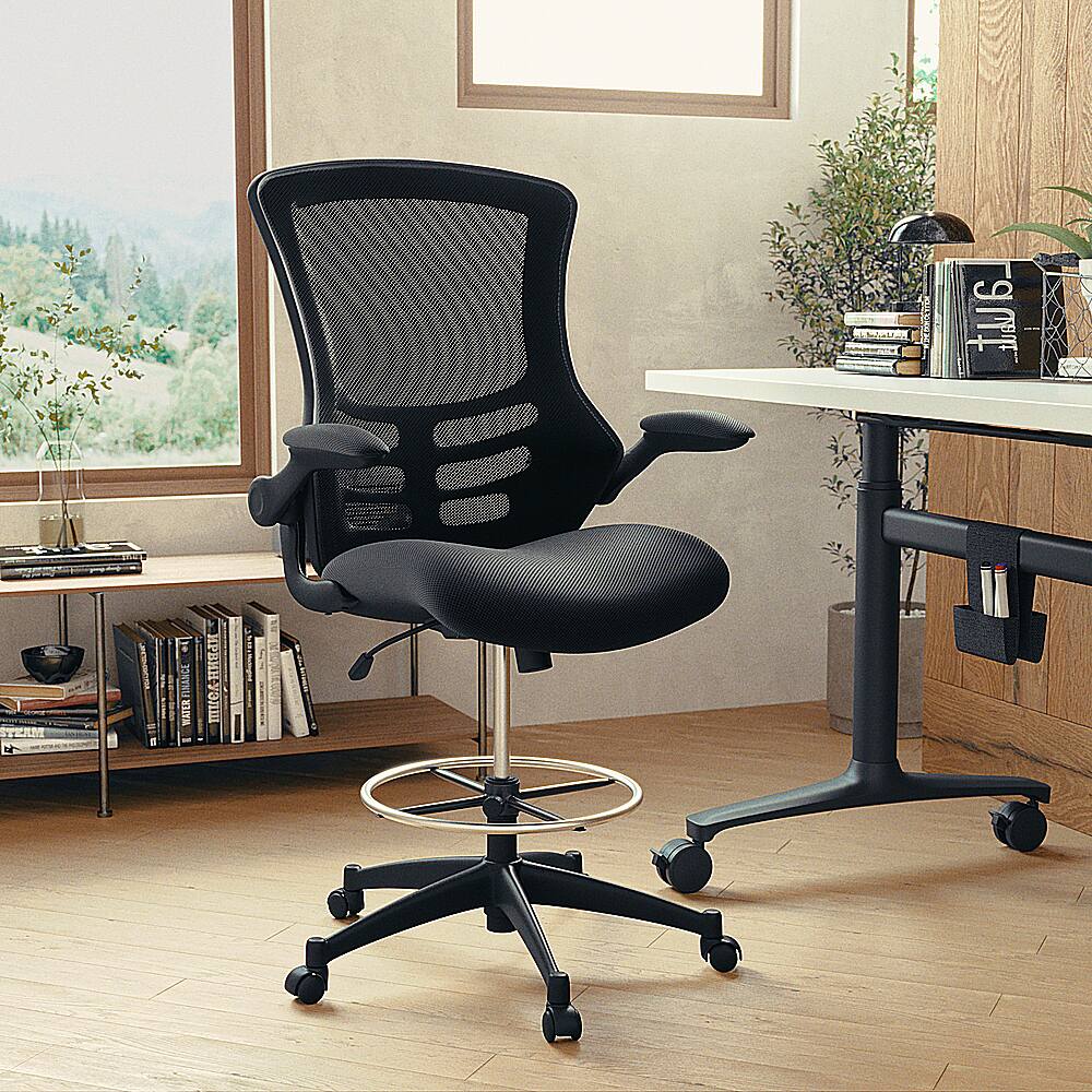 The image features a black office chair with wheels, positioned in front of a desk. The desk has a keyboard and a mouse on it. There are several books scattered around the desk, and a potted plant is placed nearby. Additionally, there is a vase on the desk, and a bottle can be seen in the background. The scene appears to be a well-organized workspace.