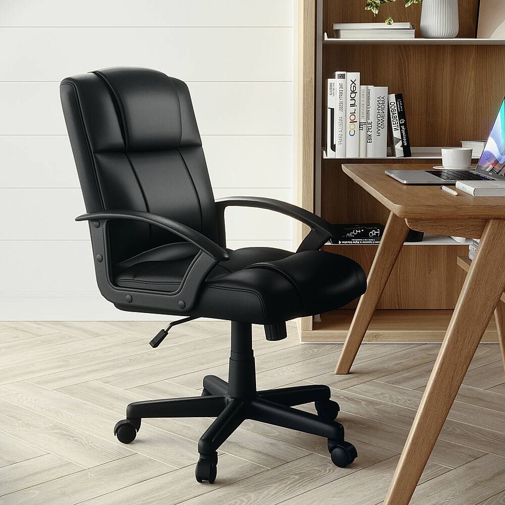 The image features a black office chair positioned in front of a wooden desk. The desk is equipped with a laptop, a keyboard, and several books. There is also a cup placed on the desk. The chair is situated in a corner of the room, and it appears to be a comfortable and functional workspace.
