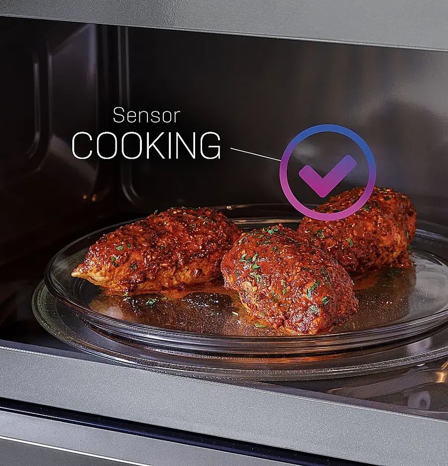The image features a microwave with a plate of food inside it. The microwave is labeled "Sensor Cooking," indicating that it uses sensors to monitor and adjust the cooking process. The plate contains three pieces of food, possibly meatballs, and is placed on a glass plate. The microwave is open, revealing the food and the cooking process taking place.