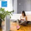 The image features a woman sitting on the floor in a bedroom, holding a cell phone. She is positioned next to a bed and a potted plant. There is a large Energy Star logo in the top left corner of the image, which suggests that the room is energy-efficient. The woman appears to be focused on her phone, possibly texting or browsing the internet.
