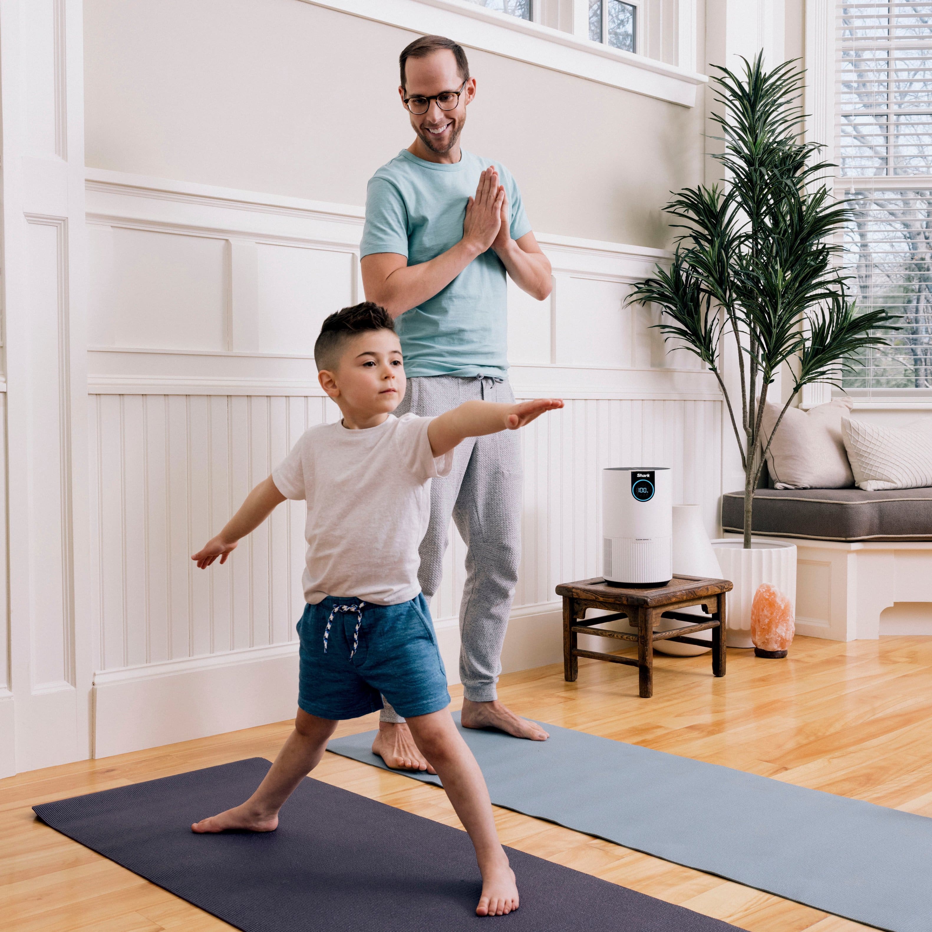 The image shows a man and a young boy standing in a living room, with the man wearing glasses and a blue shirt. They are both engaged in a fun activity, possibly playing a game. The room has a comfortable and cozy atmosphere, with a couch and a potted plant in the background. The presence of a remote control suggests that they might be using it to control a device, such as a TV or a gaming console. The scene conveys a sense of bonding and enjoyment between the man and the boy, as they spend quality time together in the living room.