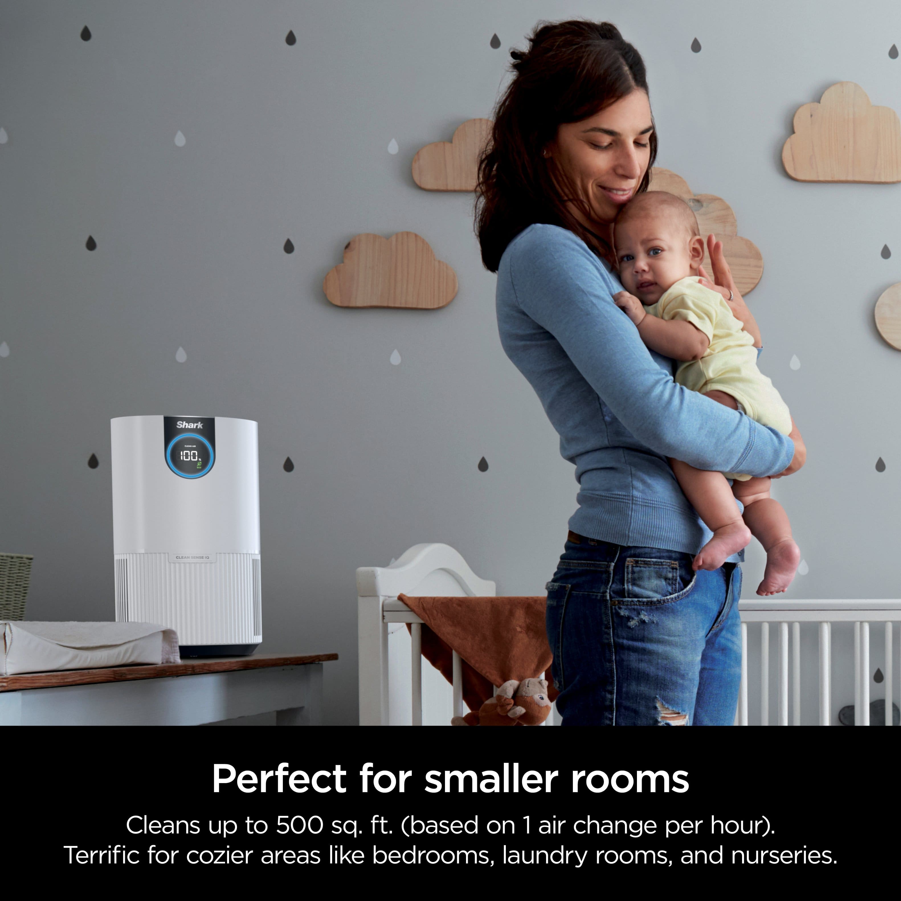 The image features a woman holding a baby while standing in a room. The room is furnished with a crib, a chair, and a dining table. The woman is wearing a blue shirt and appears to be the mother of the baby. The room is described as perfect for smaller rooms, with the ability to clean up to 500 sq. ft. based on one air change per hour. It is suitable for cozier areas like bedrooms, laundry rooms, and nurseries.