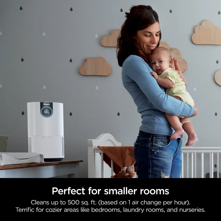 The image features a woman holding a baby while standing in a room. The room is furnished with a crib, a chair, and a dining table. The woman is wearing a blue shirt and appears to be the mother of the baby. The room is described as perfect for smaller rooms, with the ability to clean up to 500 sq. ft. based on one air change per hour. It is suitable for cozier areas like bedrooms, laundry rooms, and nurseries.