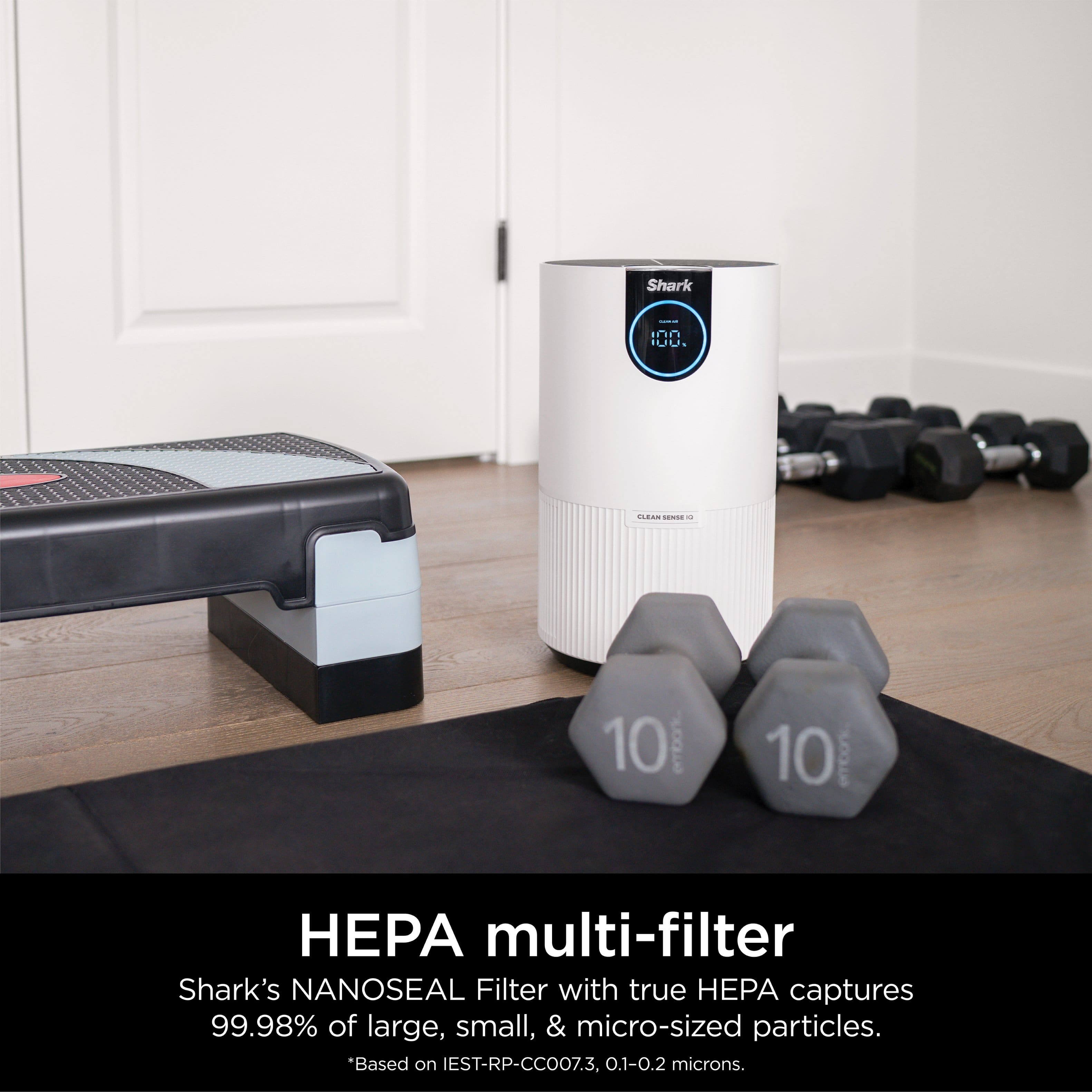The image features a white Shark HEPA multi-filter with two black dumbbells on a black mat. The filter is designed to capture 99.98% of large, small, and micro-sized particles, providing efficient air purification. The presence of the dumbbells suggests that the scene might be set in a gym or a workout area.