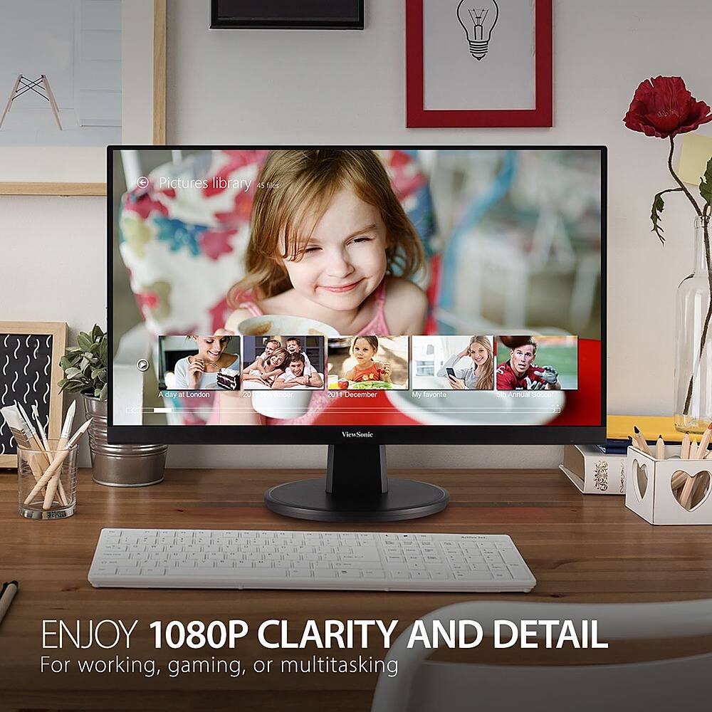 The image features a computer monitor displaying a photo of a young girl. The monitor is placed on a desk, surrounded by various items such as a keyboard, a mouse, and a vase. There are also several books scattered around the desk, and a potted plant is located nearby. The scene suggests a workspace or a home office setup.
