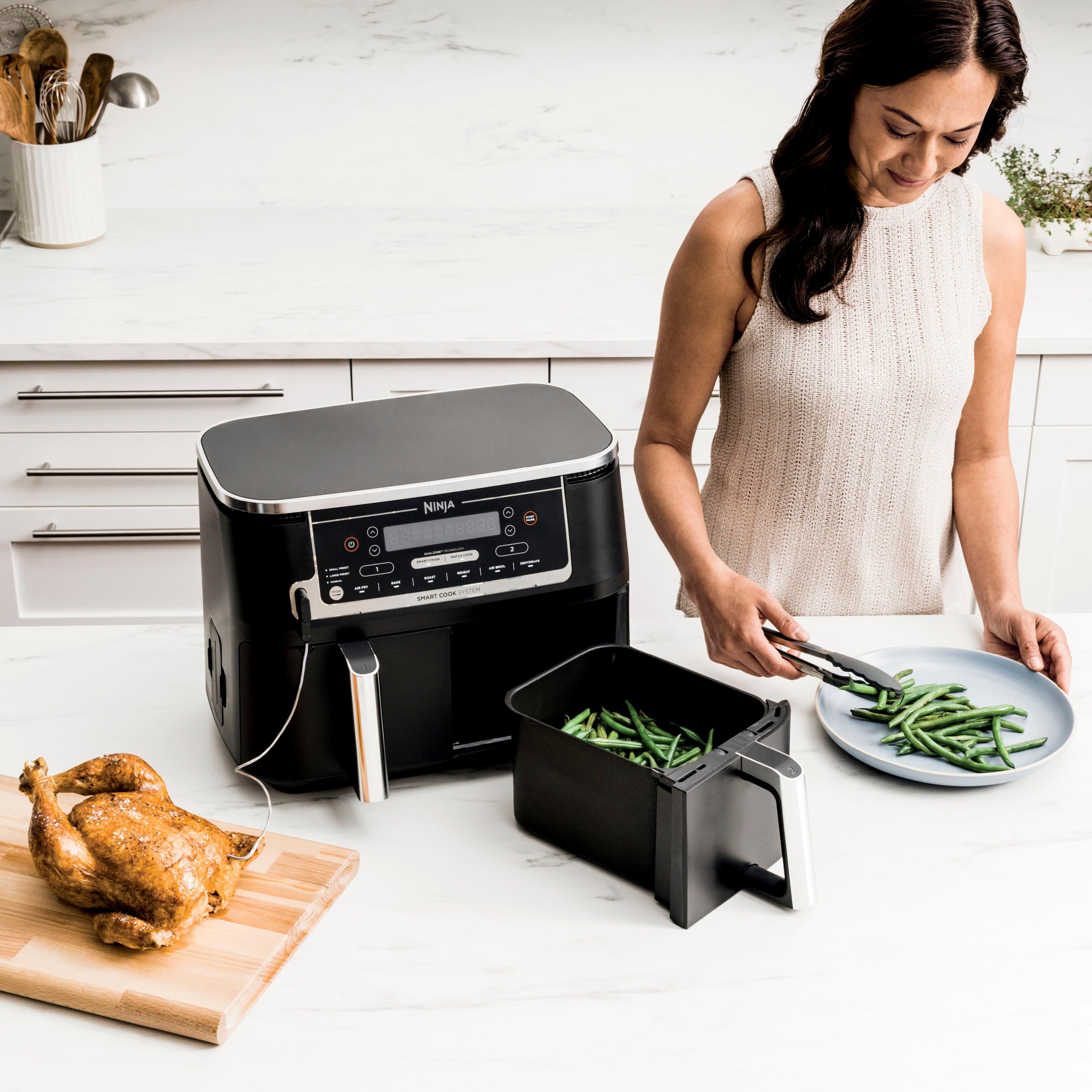 The image features a chicken and a vegetable dish, with the chicken placed on a cutting board and the vegetables in a black bowl. The dish is being prepared using a Ninja food system, which is a smart appliance that allows for efficient cooking and finishing of both mains and sides simultaneously. The image promotes the convenience and efficiency of using the Ninja food system for meal preparation.