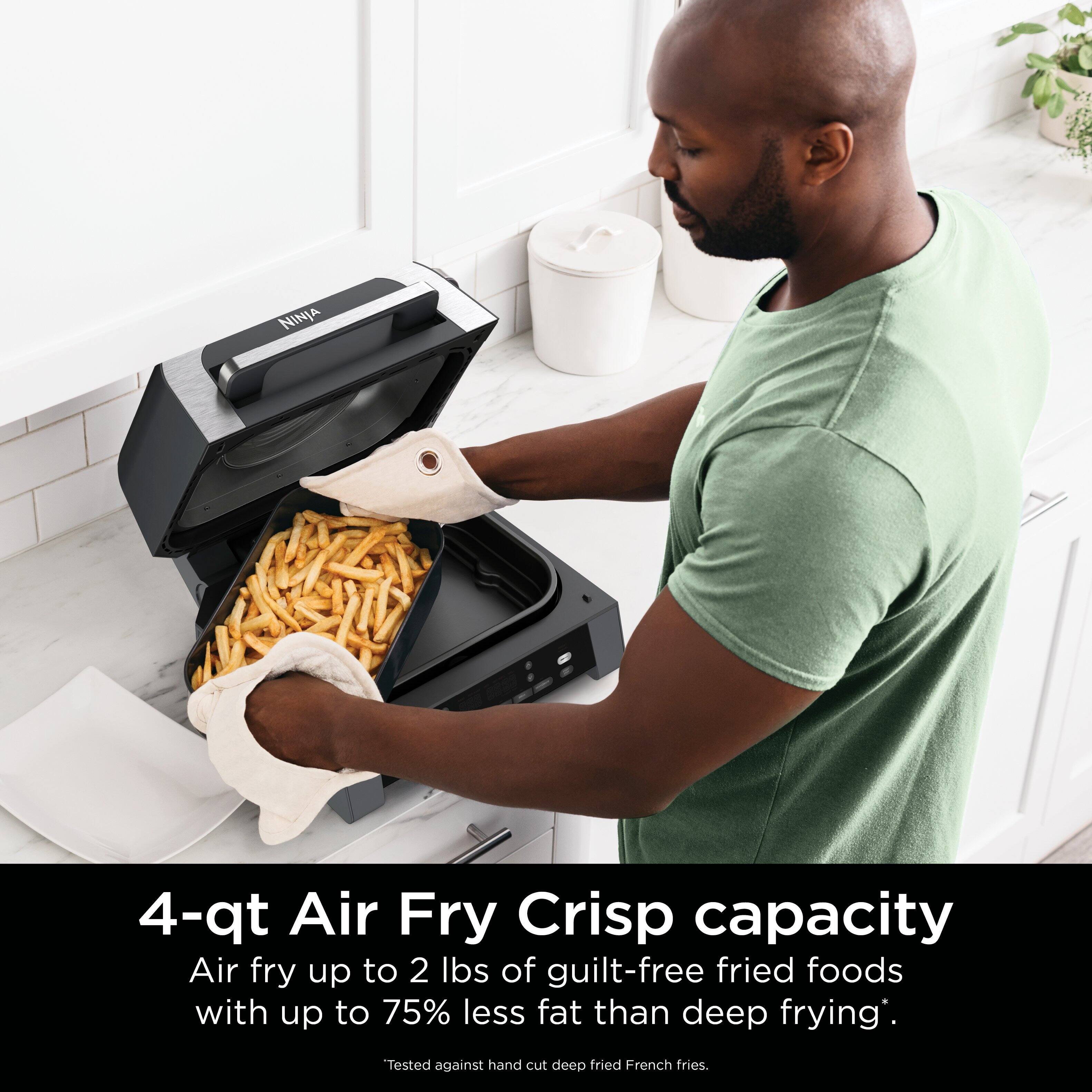 The image features a man using a Ninja 4-qt Air Fry Crisp, which has a capacity to air fry up to 2 lbs of guilt-free fried foods with up to 75% less fat than deep frying. The man is reaching into the fryer to remove the fries, which are being cooked to a crispy texture. The image also includes a potted plant in the background, adding a touch of greenery to the scene.