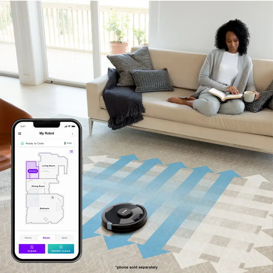 The image features a woman sitting on a couch, holding a book and a cell phone. The cell phone is displaying a map of the house, possibly indicating that a robotic vacuum is being used to clean the house. The woman is sitting on a beige couch, and there are multiple potted plants in the room. A cup can also be seen on a surface nearby.