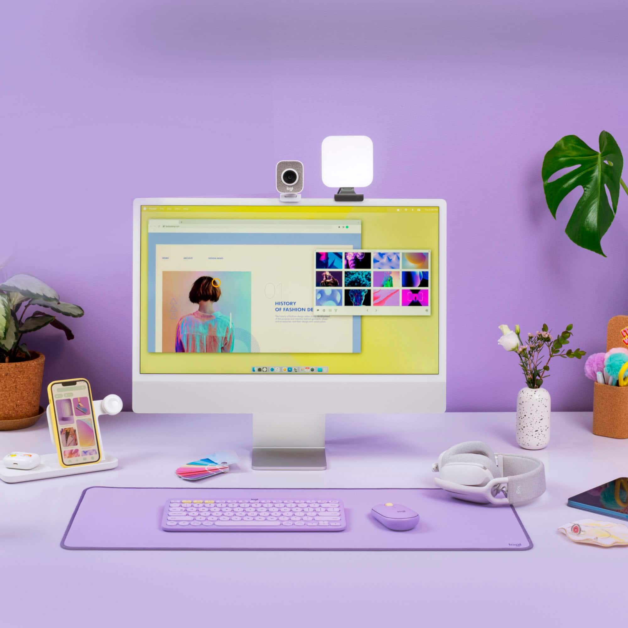 The image features a computer desk with a desktop computer, keyboard, mouse, and a cell phone. There are also several books and a potted plant on the desk. The computer is turned on, and the screen displays a woman's face. The keyboard is purple, and there are two cell phones on the desk. Additionally, there are two vases and a potted plant nearby. The scene appears to be a workspace or a home office.