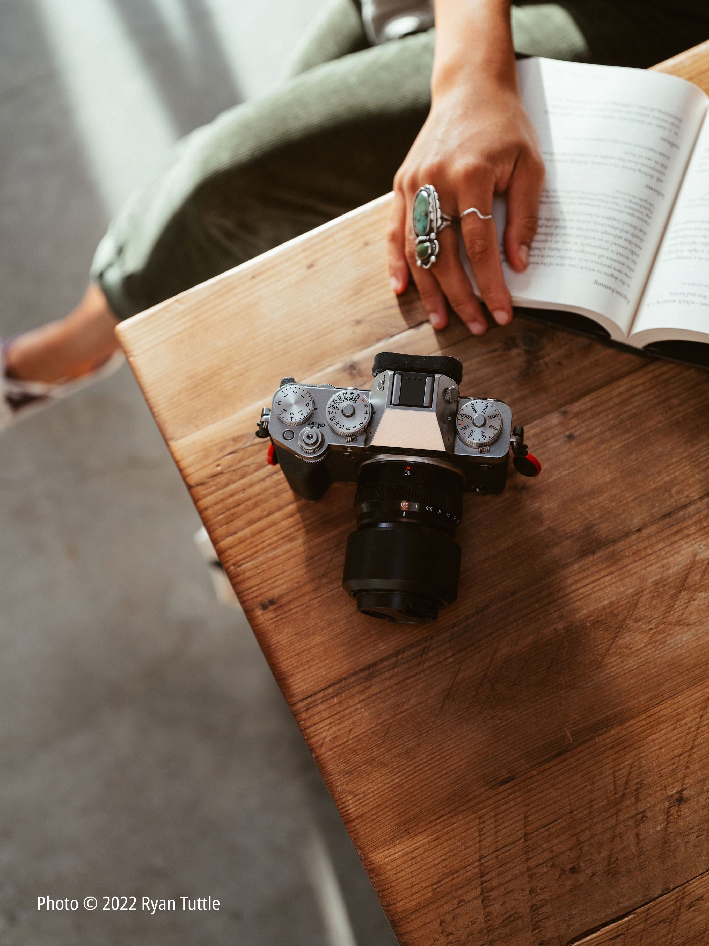 The image features a person sitting at a table with a camera and a book. The person is holding the camera in their hand, and there is a book placed nearby. The scene appears to be a casual setting where the person is likely taking a break from reading or working to engage with the camera. The presence of the camera suggests that the person might be interested in photography or capturing memories.