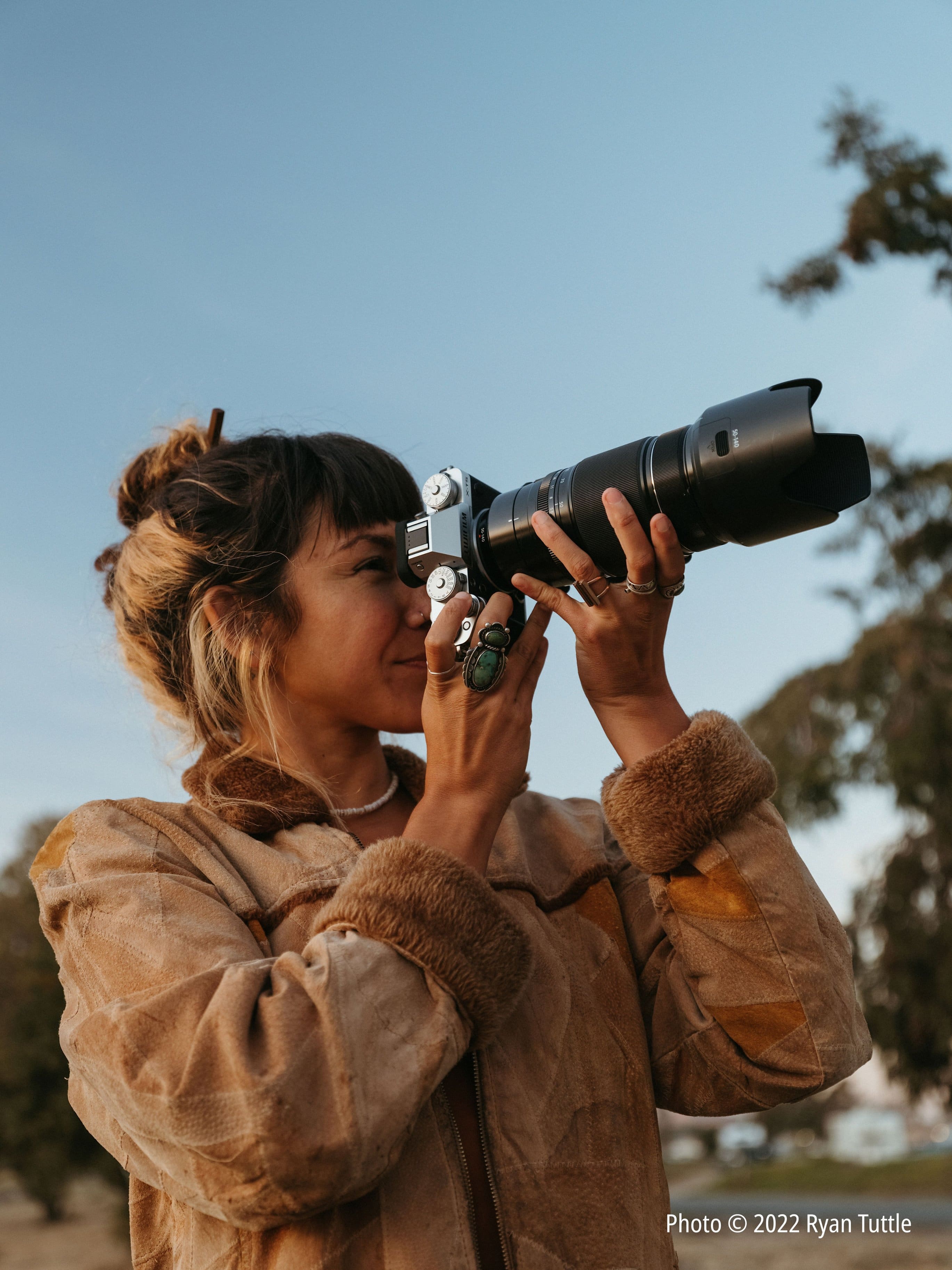 The image features a woman wearing a brown jacket and holding a camera up to her eye. She appears to be taking a picture, possibly of the sky. The woman is the main focus of the image, and her camera is prominently visible. The scene is likely related to photography or capturing moments in nature, as the woman is looking through the camera lens.