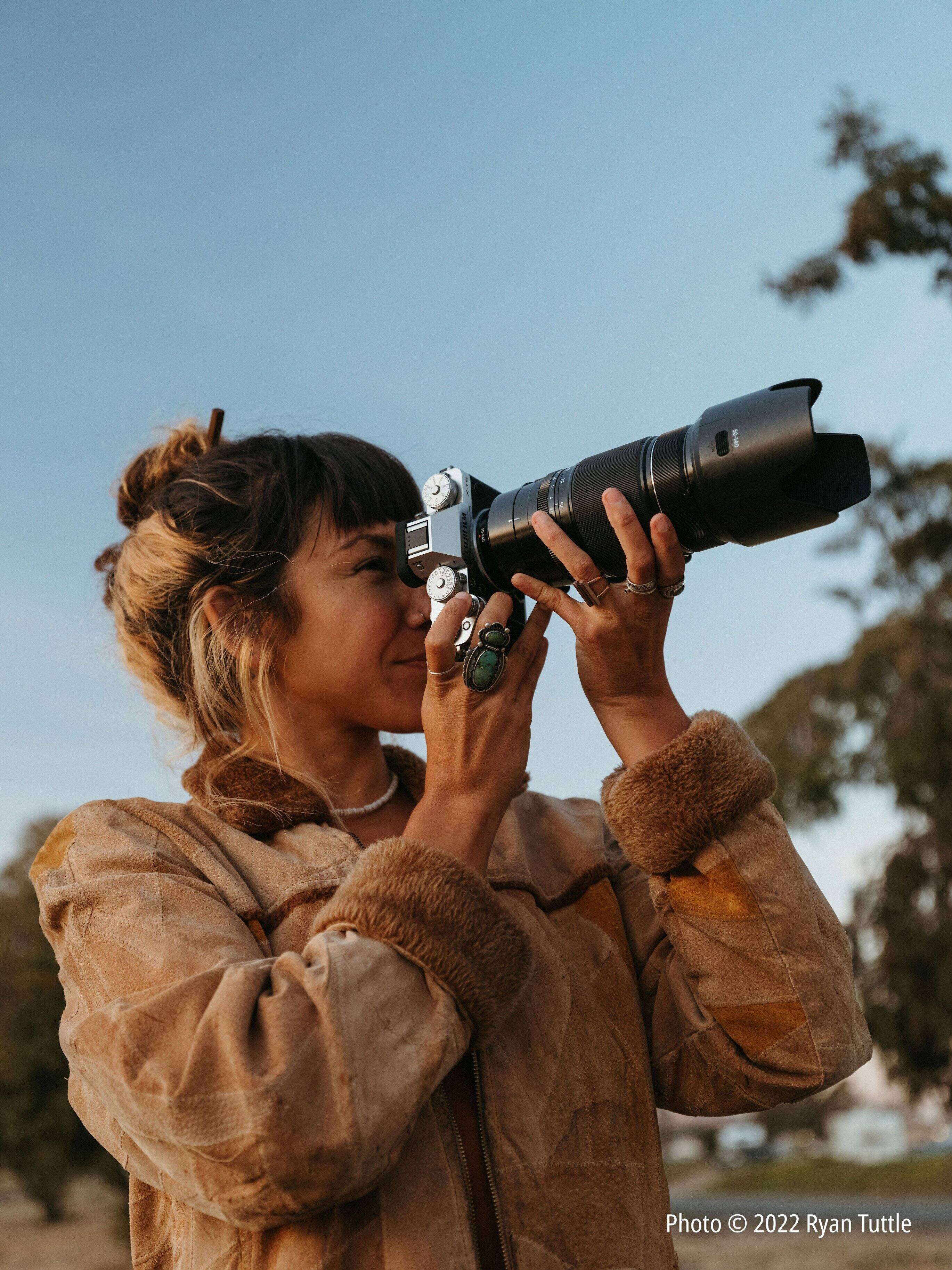 The image features a woman wearing a brown jacket and holding a camera up to her eye. She appears to be taking a picture, possibly of the sky. The woman is the main focus of the image, and her camera is prominently visible. The scene is likely related to photography or capturing moments in nature, as the woman is looking through the camera lens.