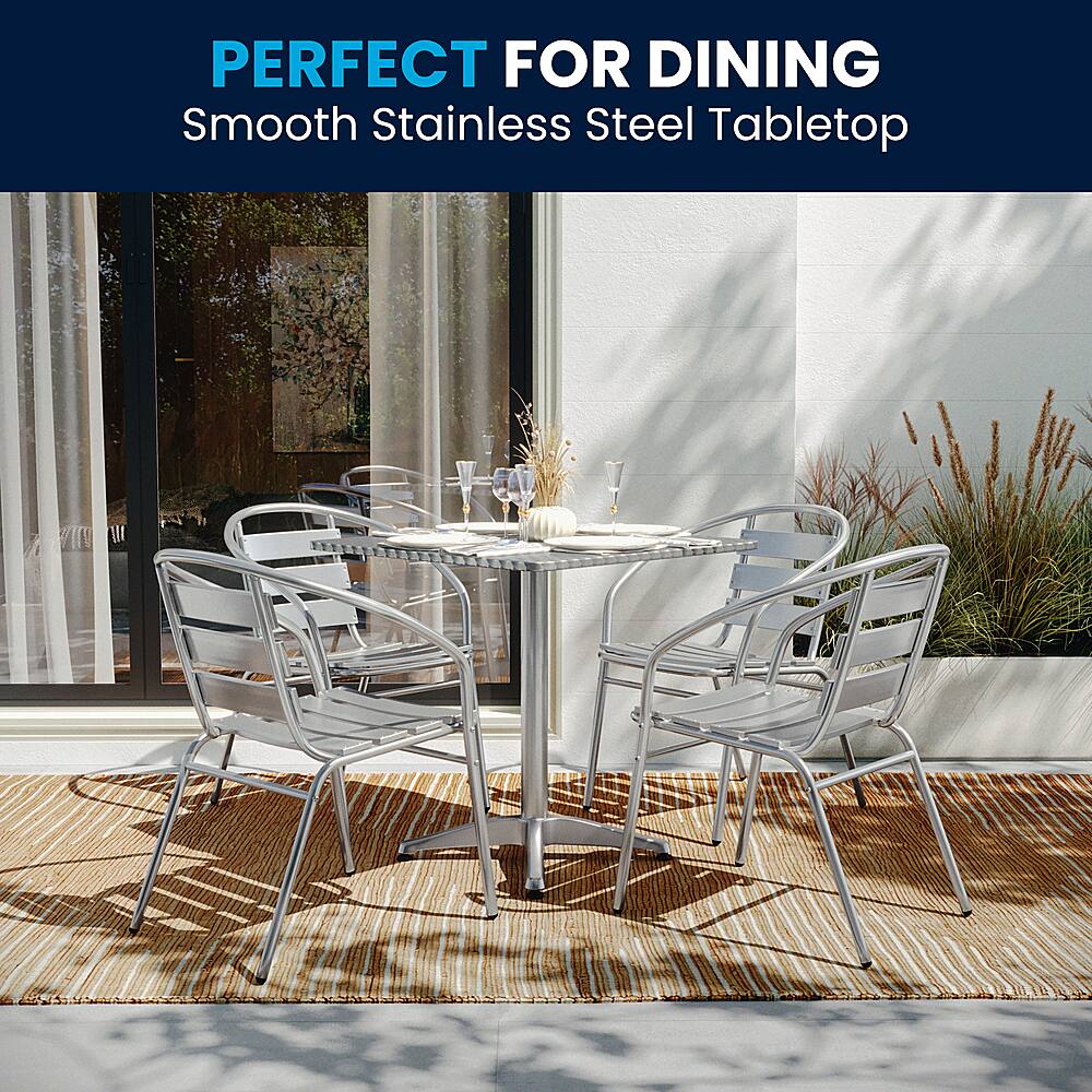 The image showcases a dining table with four chairs around it, placed on a smooth stainless steel tabletop. The table is set with various items, including wine glasses, cups, and a vase. The table is surrounded by a few potted plants, adding a touch of greenery to the scene. The chairs are arranged in a way that encourages conversation and socializing, making it an ideal setup for a pleasant dining experience.