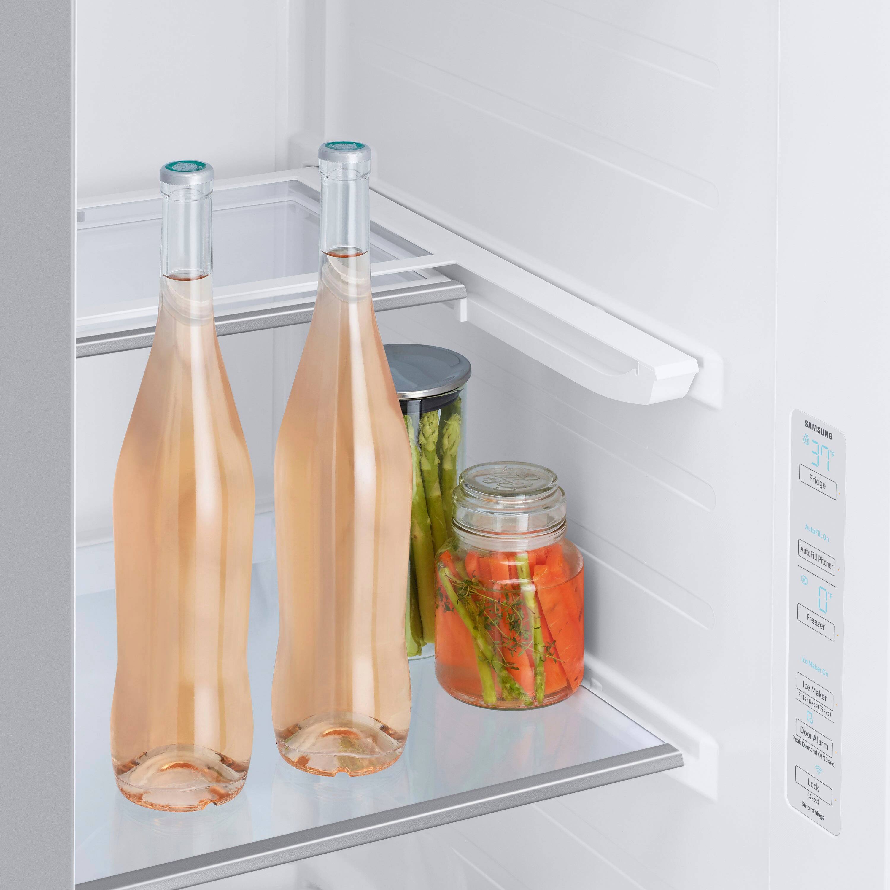 The image shows a refrigerator with two bottles of wine and a pickle jar inside. The refrigerator has a door alarm, which is designed to alert users if the door is left open. The wine bottles are placed on the refrigerator shelf, while the pickle jar is located on the door shelf. The refrigerator is white, and the bottles and jar are placed in the middle of the refrigerator.