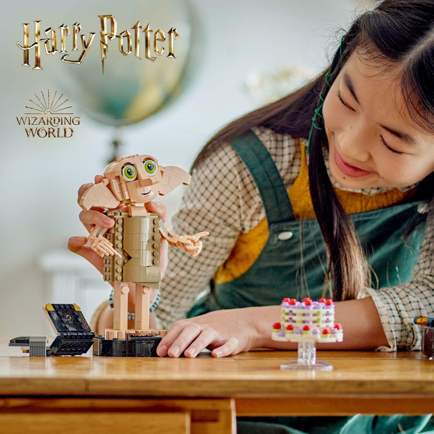 The image features a young girl playing with a Harry Potter toy, which is a part of the Wizarding World. She is sitting at a table, holding the toy and smiling. The toy appears to be a small figurine or a building set, adding to the excitement and fun of the playtime.