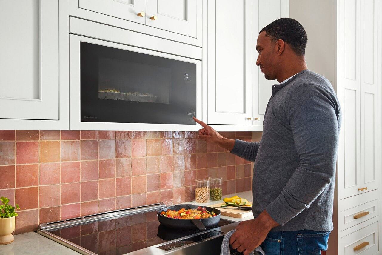 The image features a person holding a microwave door open, with the microwave placed above a stove. The microwave is white and appears to be a KitchenAid model. The person is likely in the process of installing or adjusting the microwave door.