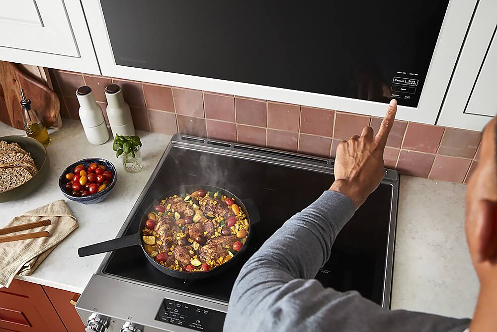 The image features a man cooking food in a pan on a stove. He is pointing at the food, indicating that he is in the process of preparing a meal. The scene takes place in a kitchen, with a microwave visible in the background. The microwave is placed on the counter, and it is not in use at the moment. The man's focus is on the food he is cooking, and he is likely enjoying the process of creating a delicious meal.