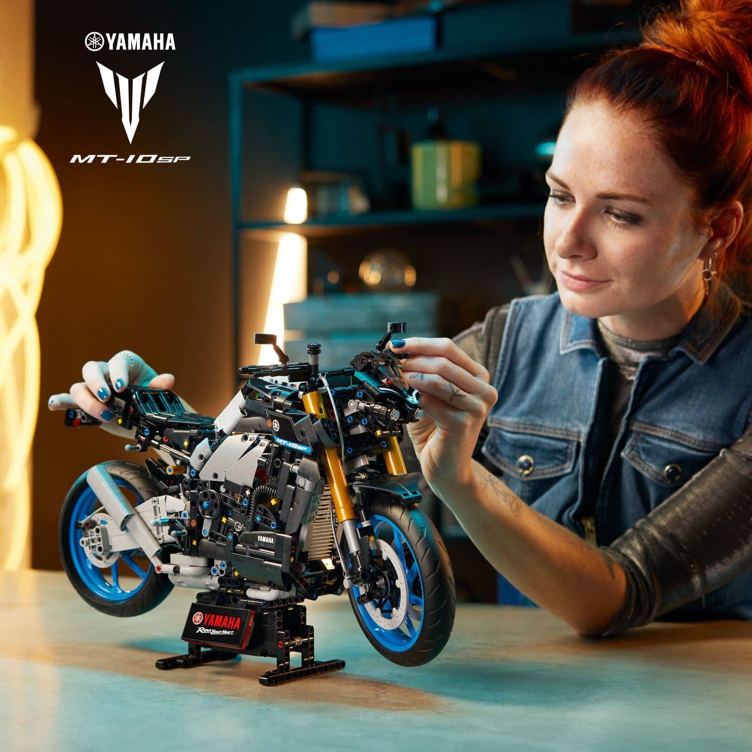 The image features a woman sitting at a table, assembling a toy motorcycle made of Legos. She is carefully putting the pieces together, creating a detailed and intricate model of a Yamaha motorcycle. The table is filled with various Lego pieces, and the woman appears to be focused on her task. The scene showcases the creativity and skill involved in constructing such a model, as well as the enjoyment that comes from building and assembling toys.